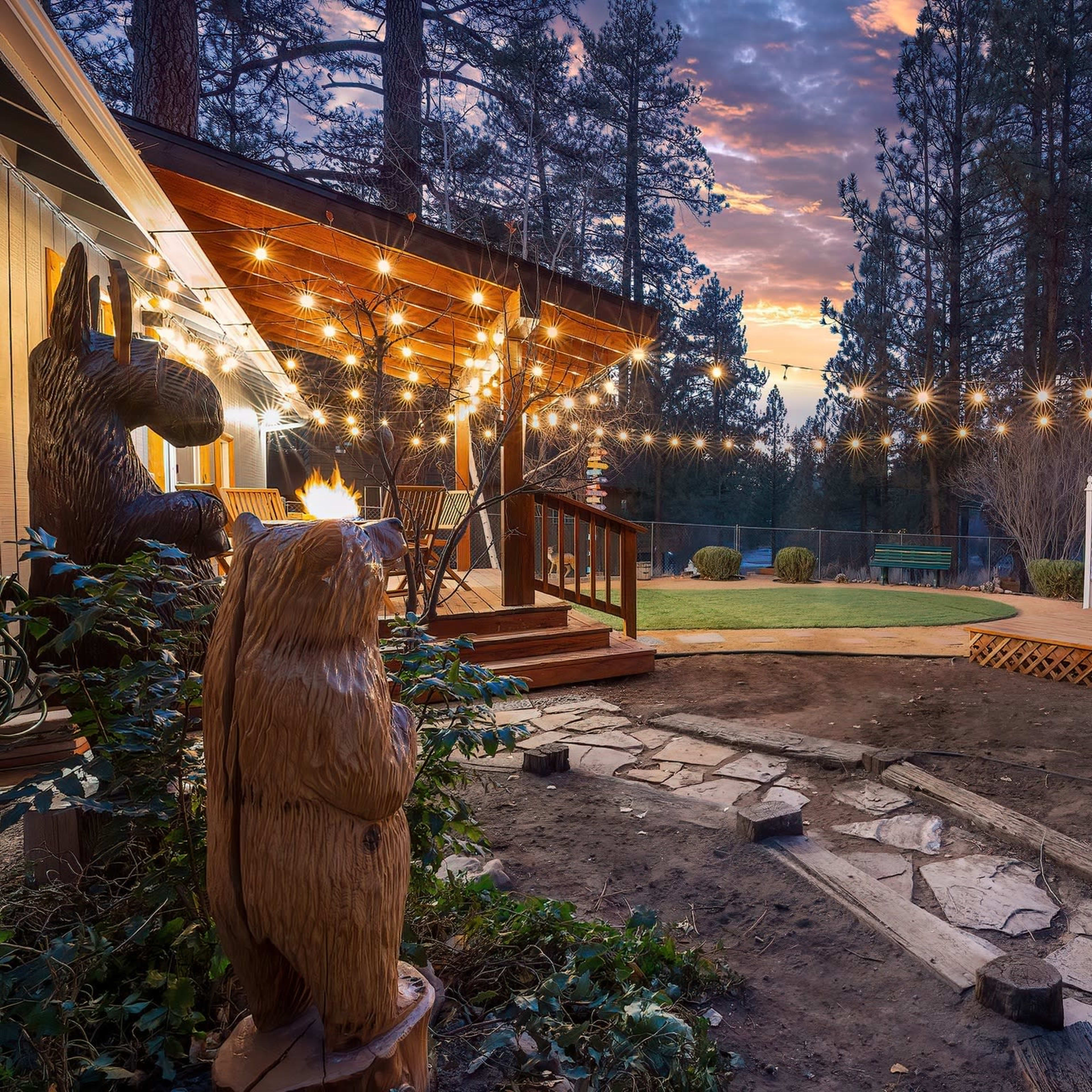 A wooden deck with string lights overlooks a landscaped yard featuring a hot tub and a bear sculpture amidst tall pine trees at sunset.