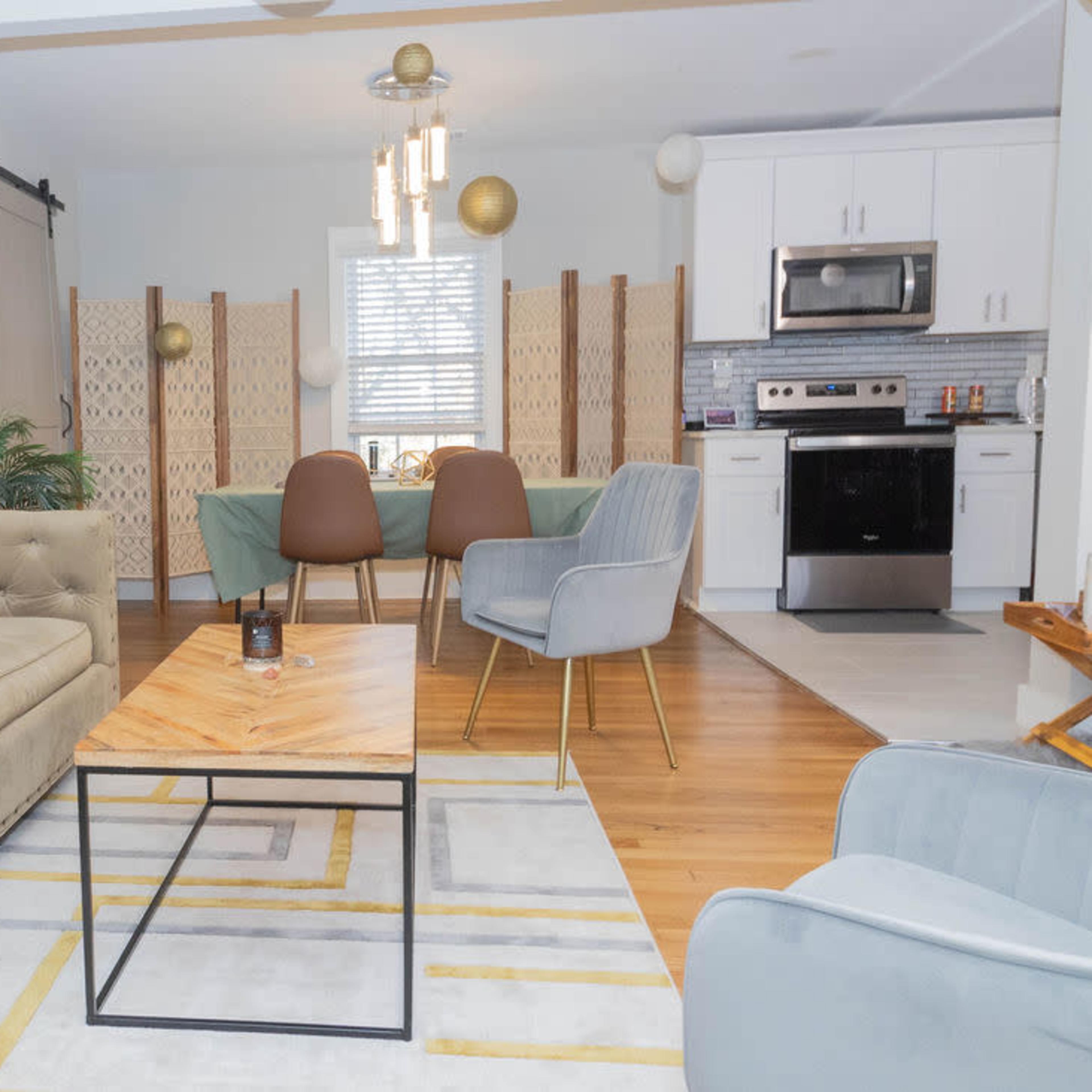 A modern living space featuring a beige couch, a wooden coffee table, and a dining area with a green tablecloth, surrounded by decorative screens and kitchen appliances.