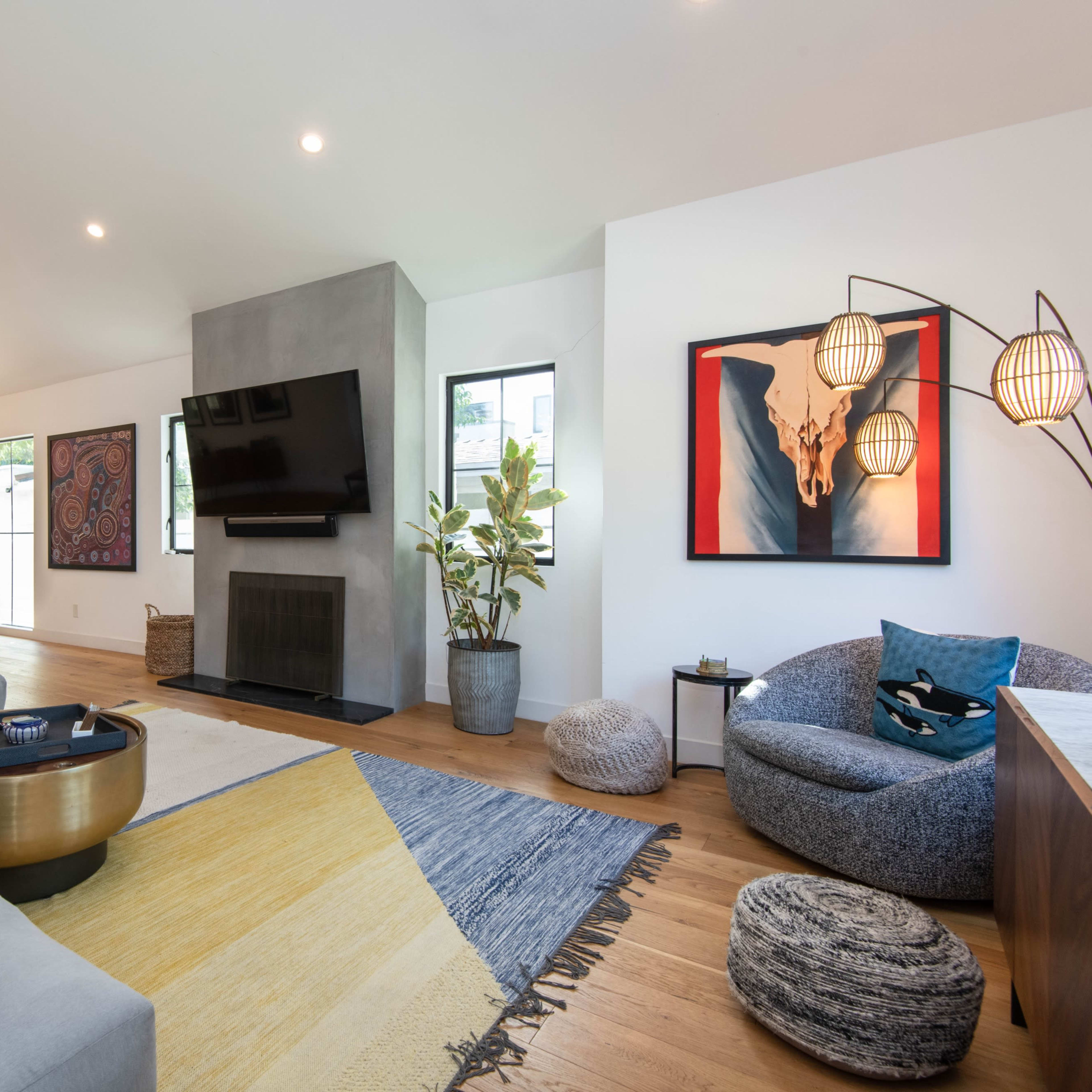 A modern living room features a sectional sofa, a large flat-screen TV mounted on a gray wall, a circular coffee table, and various decorative elements including artwork and potted plants.