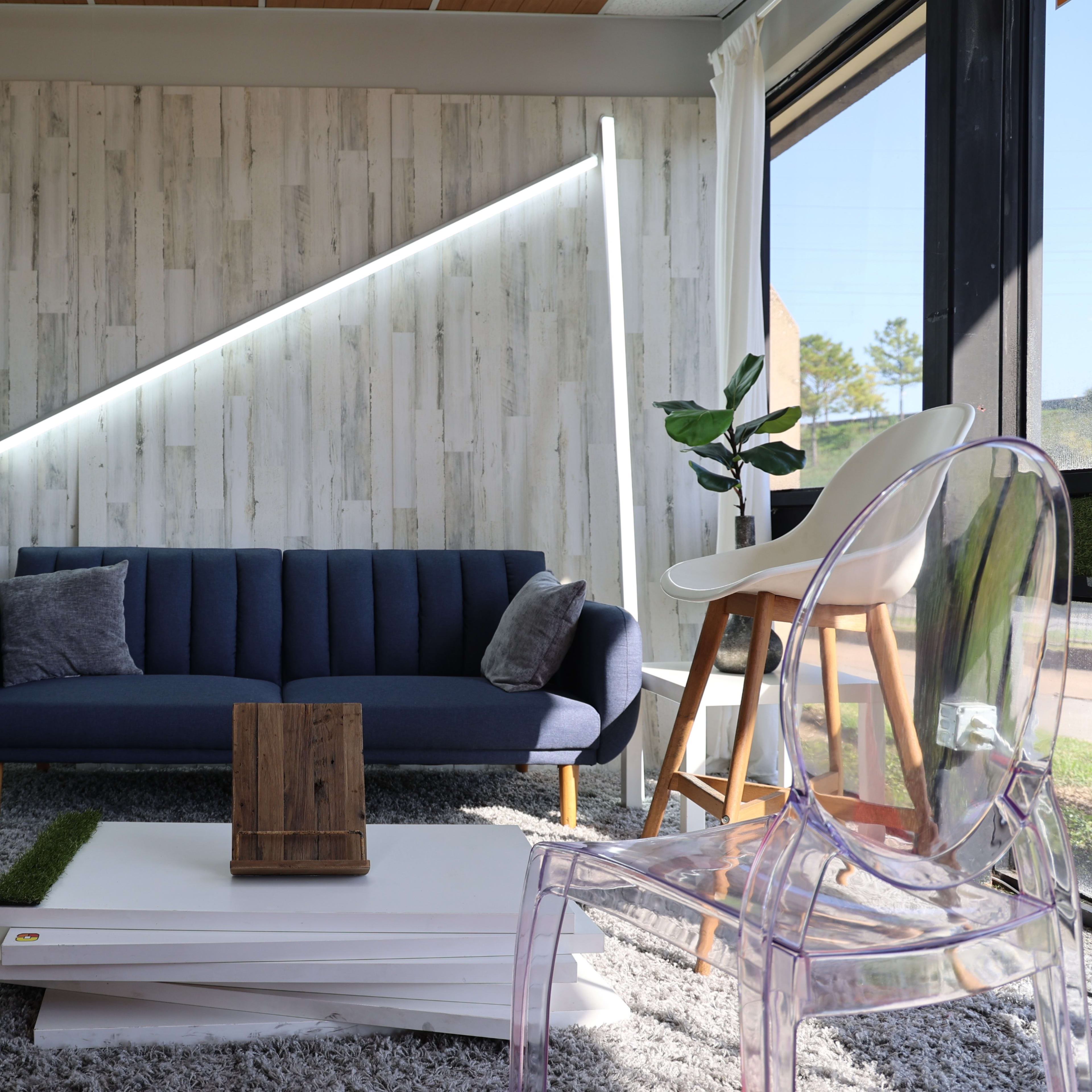 A modern living room features a blue sofa, clear chairs, and a wooden table, with plants and angled lighting against a textured wall.