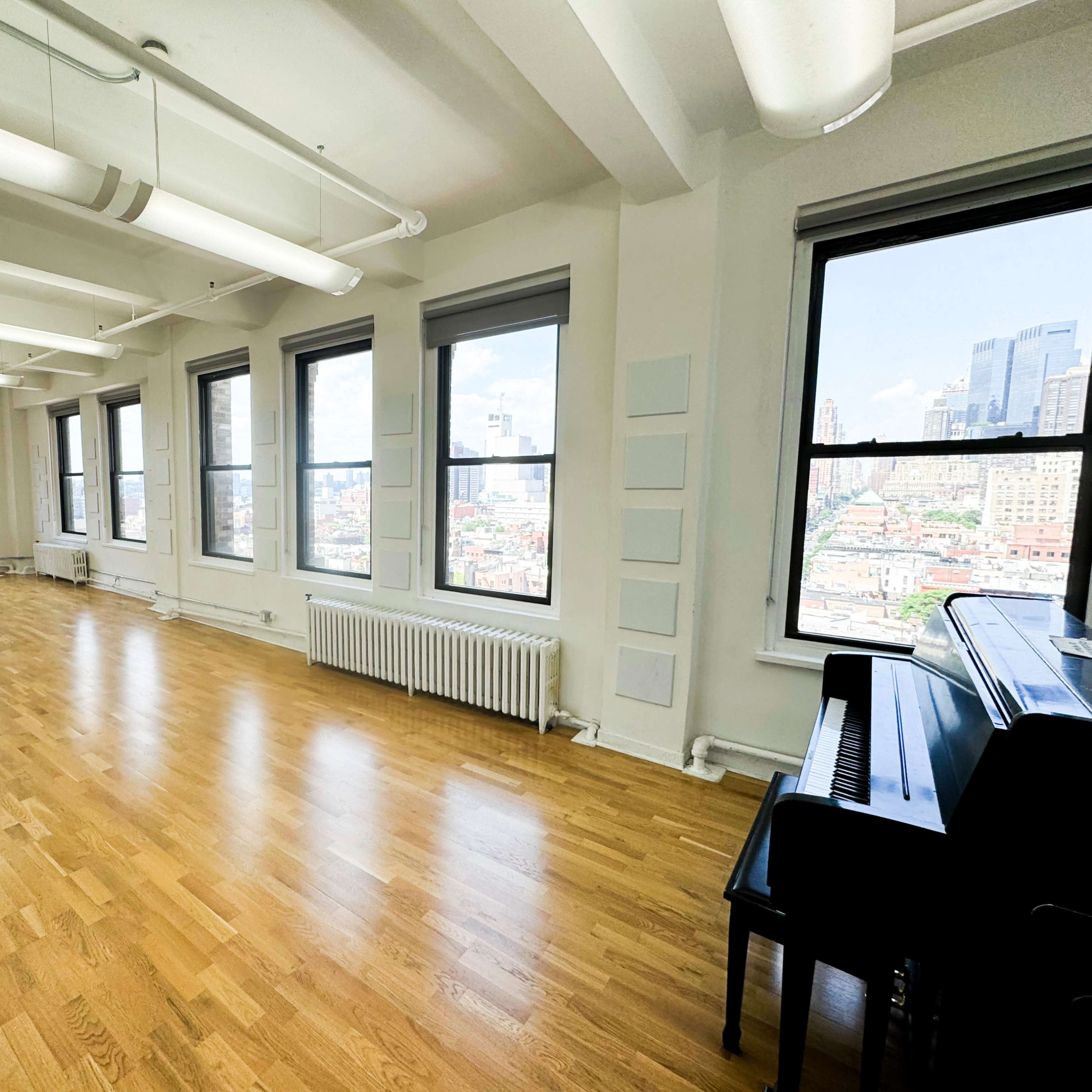 A bright, empty room with hardwood floors and large windows overlooks a cityscape, featuring a black piano against one wall.