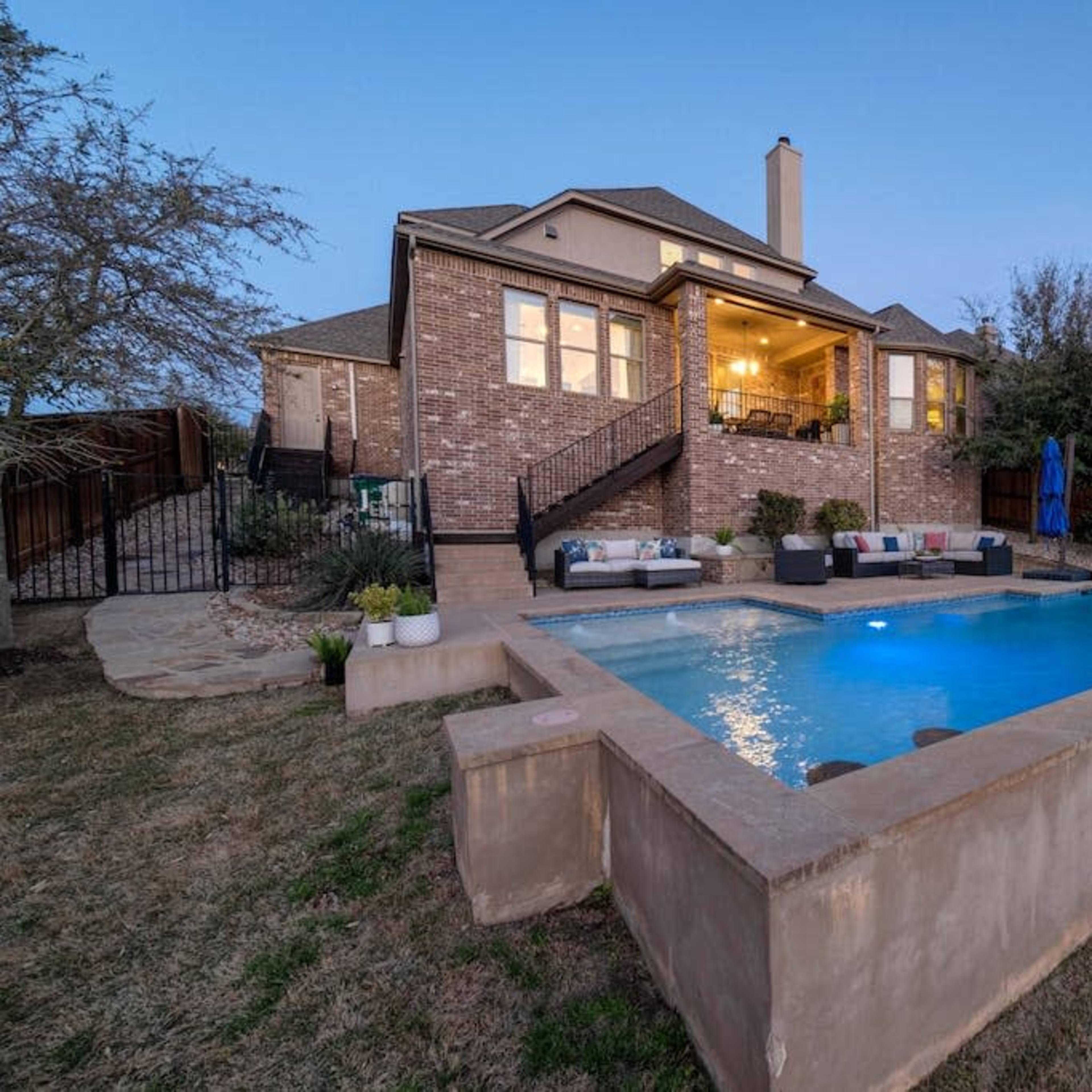 A backyard features a rectangular swimming pool with a fountain, surrounded by grass and stone landscaping, alongside a two-story brick house with outdoor seating.