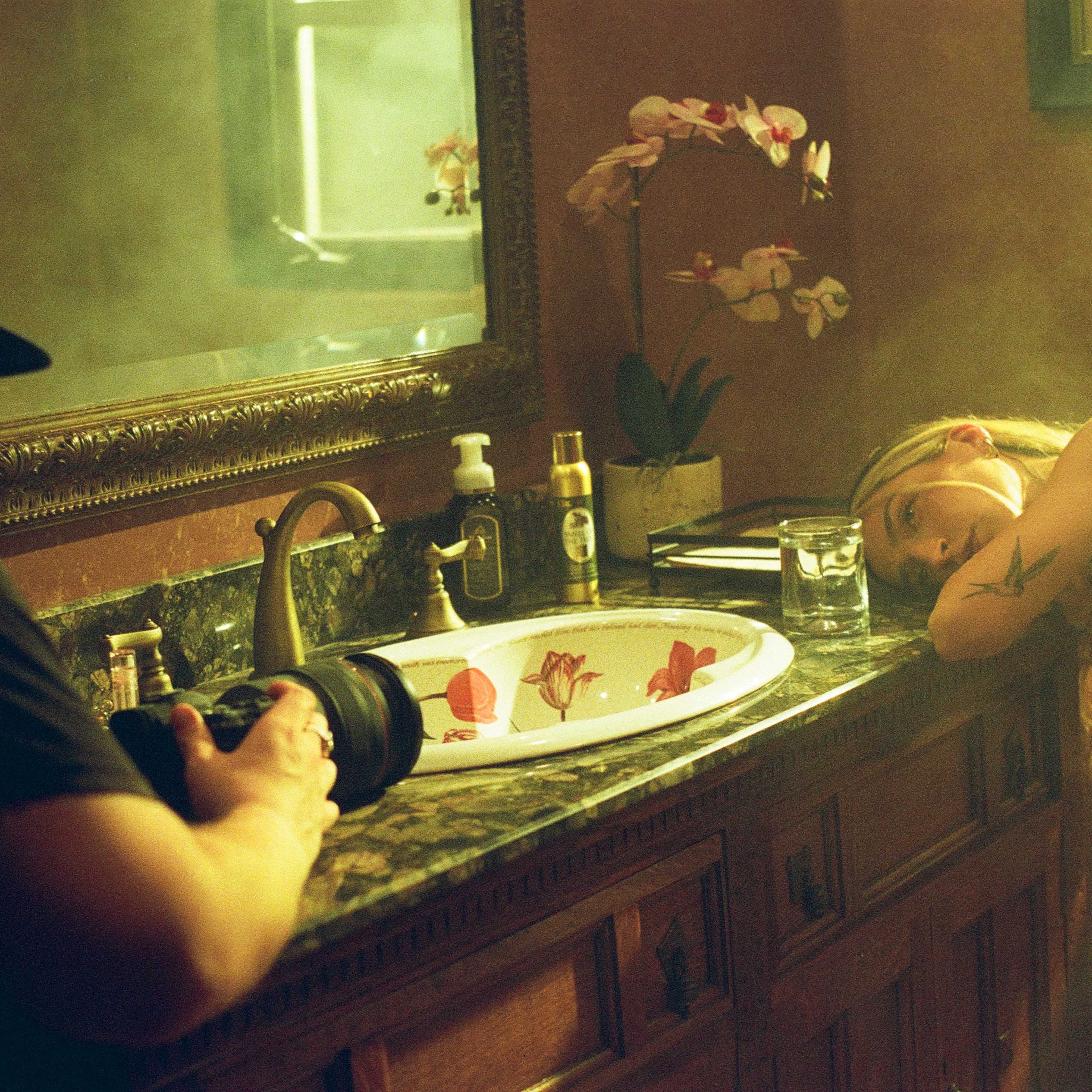 A photographer captures an image of a woman resting her head on a bathroom counter next to a sink adorned with a floral-patterned bowl.