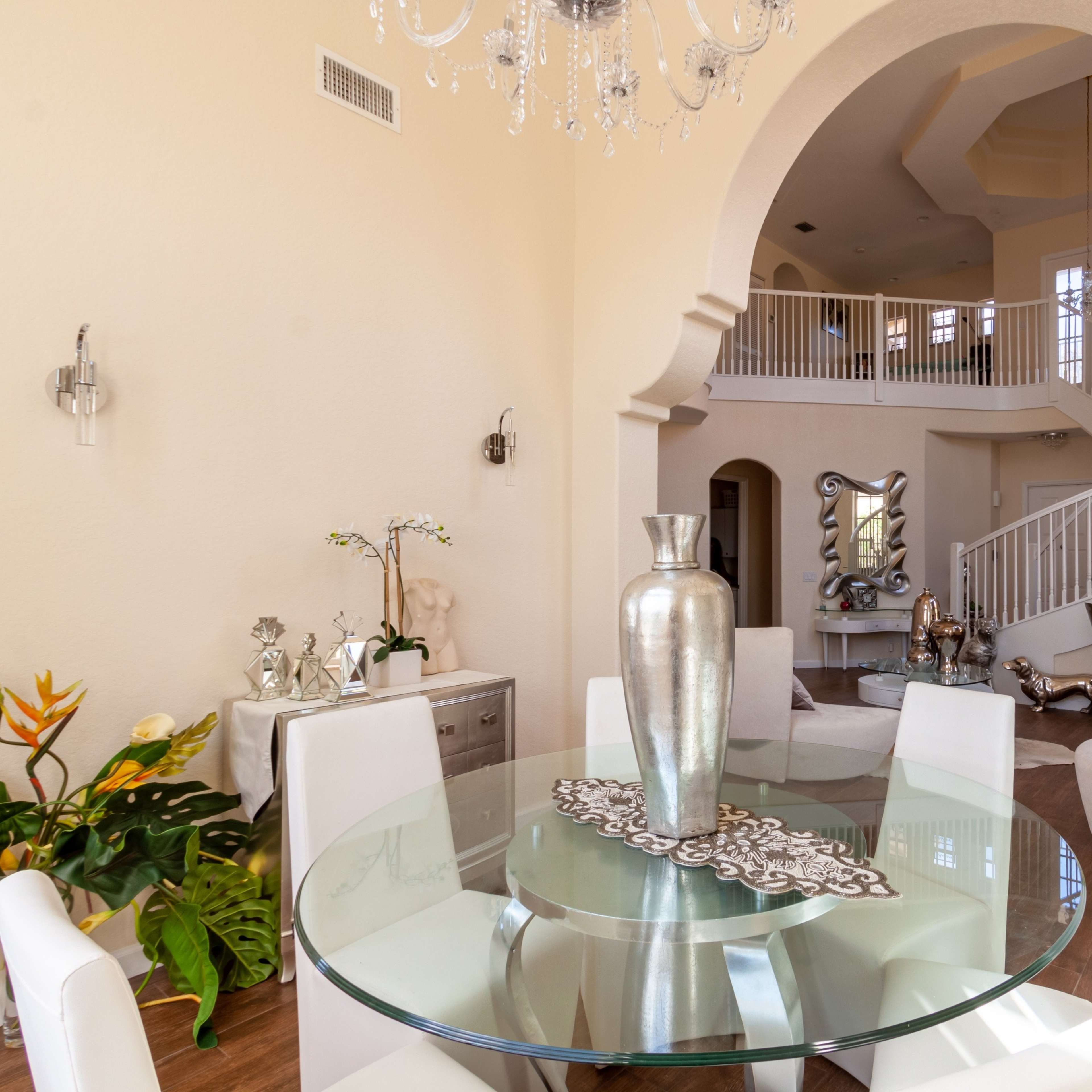 A spacious dining area with a round glass table surrounded by white chairs, leading to an elegantly designed staircase and a bar area in a bright, open-concept living space.