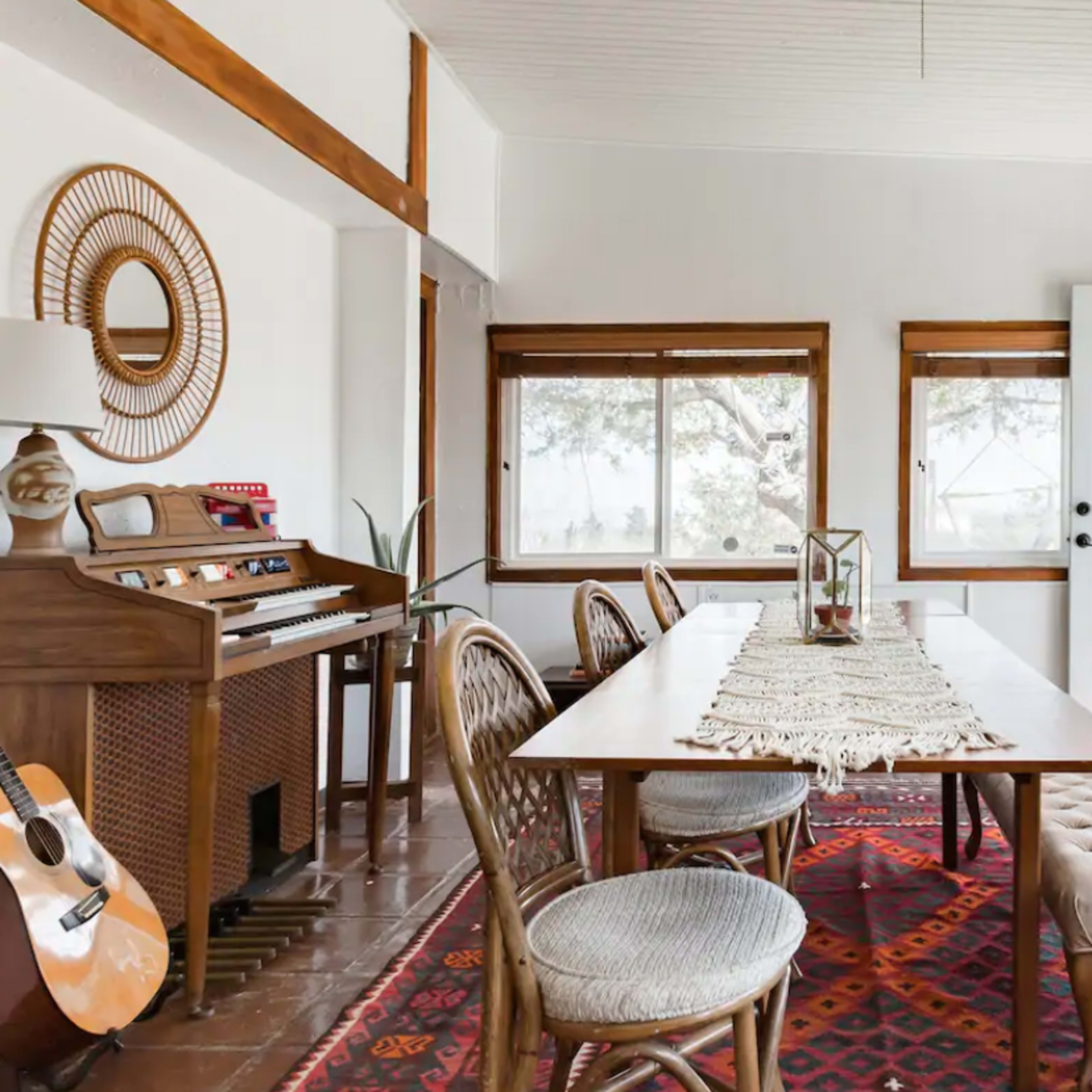 A bright room with a wooden dining table, chairs, a vintage keyboard, and large windows overlooking a scenic view.