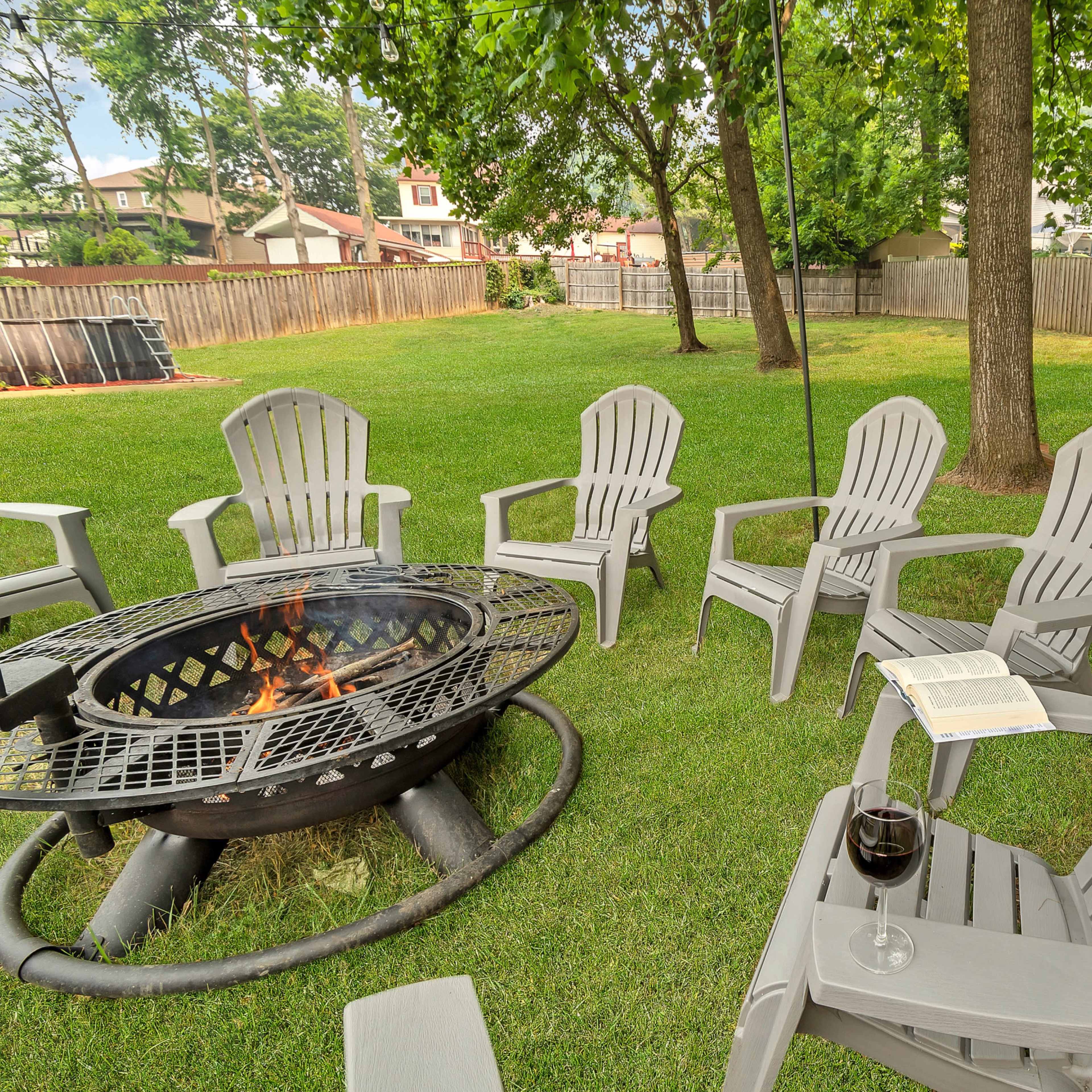 A circular fire pit with several gray chairs and a glass of wine sits on a grassy backyard lawn surrounded by trees.