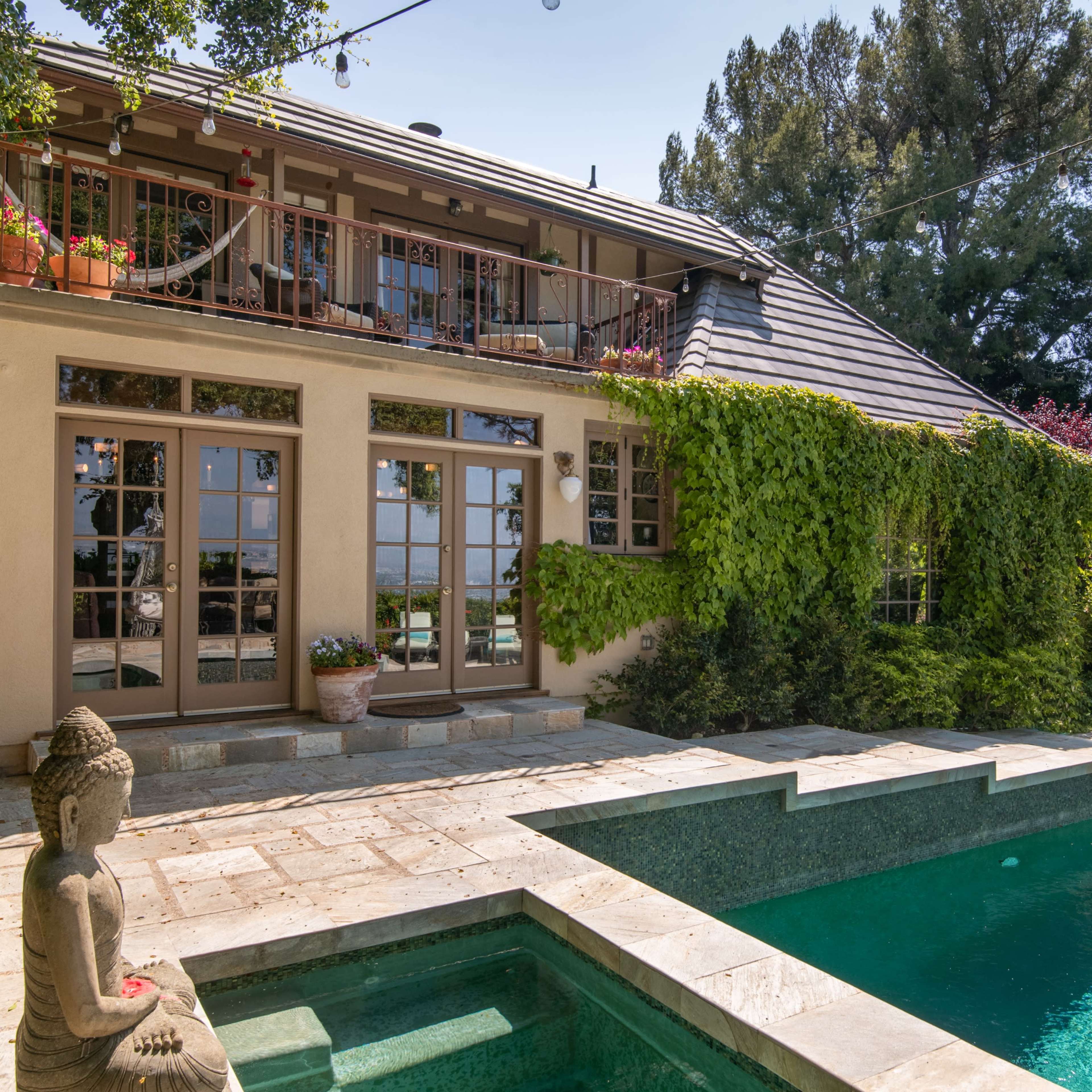 A two-story house with large windows, surrounded by greenery, next to a swimming pool with a Buddha statue at the edge.