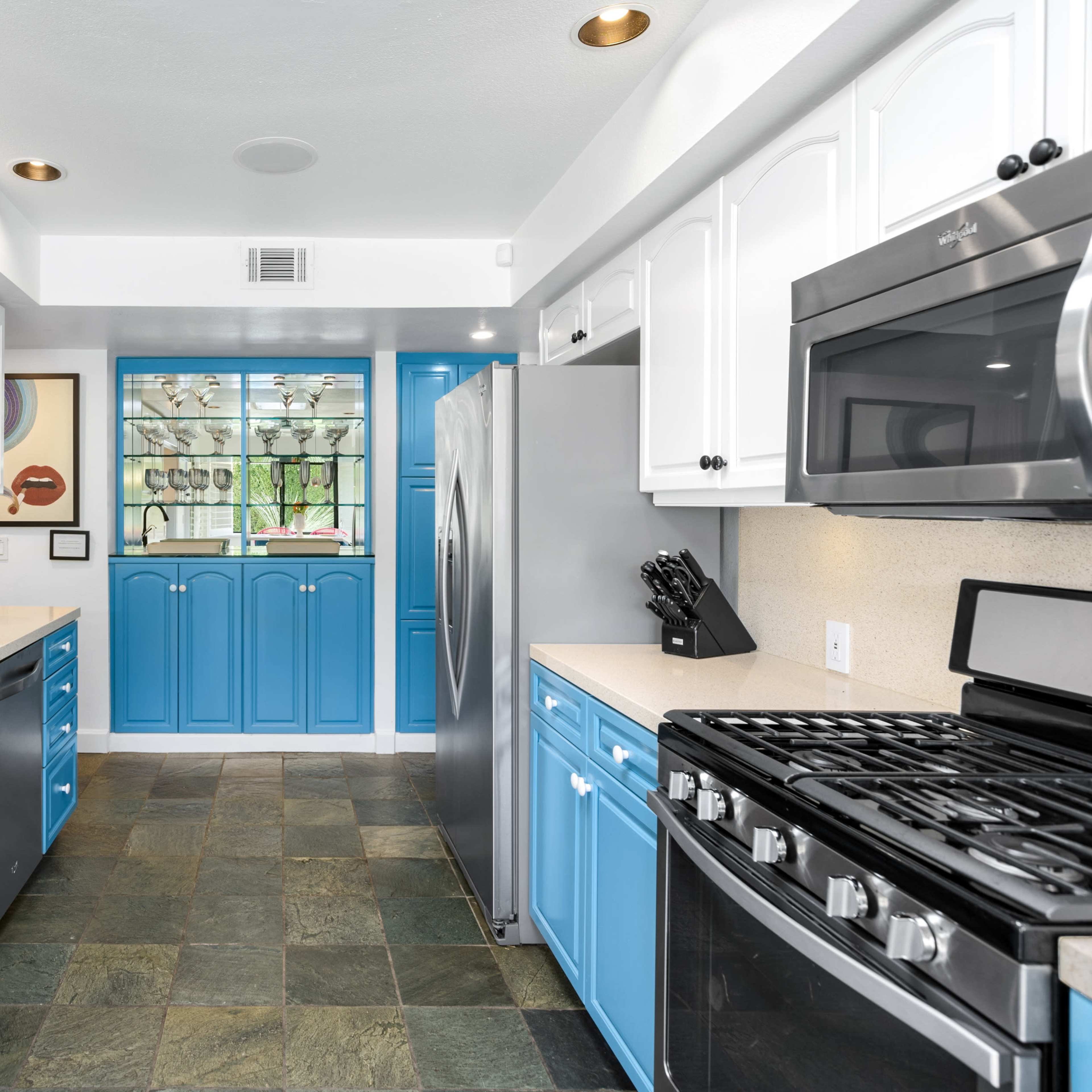 A modern kitchen with blue cabinets, a stainless steel refrigerator, and a gas stove, set against a light-colored countertop and slate flooring.