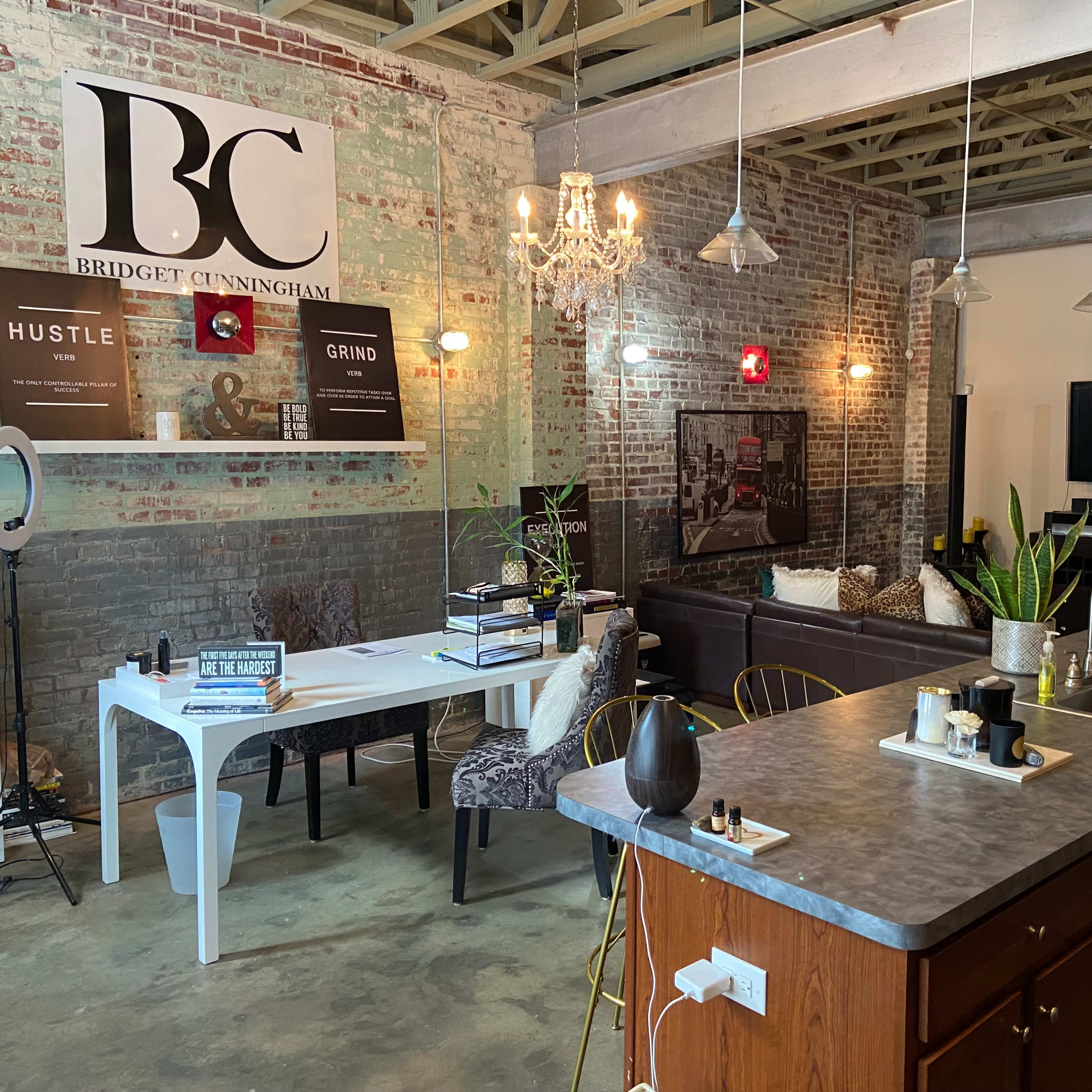 A modern interior space featuring a kitchen island with bar stools, a dining area with a white table, and a lounge area with a sofa, all set against exposed brick walls.