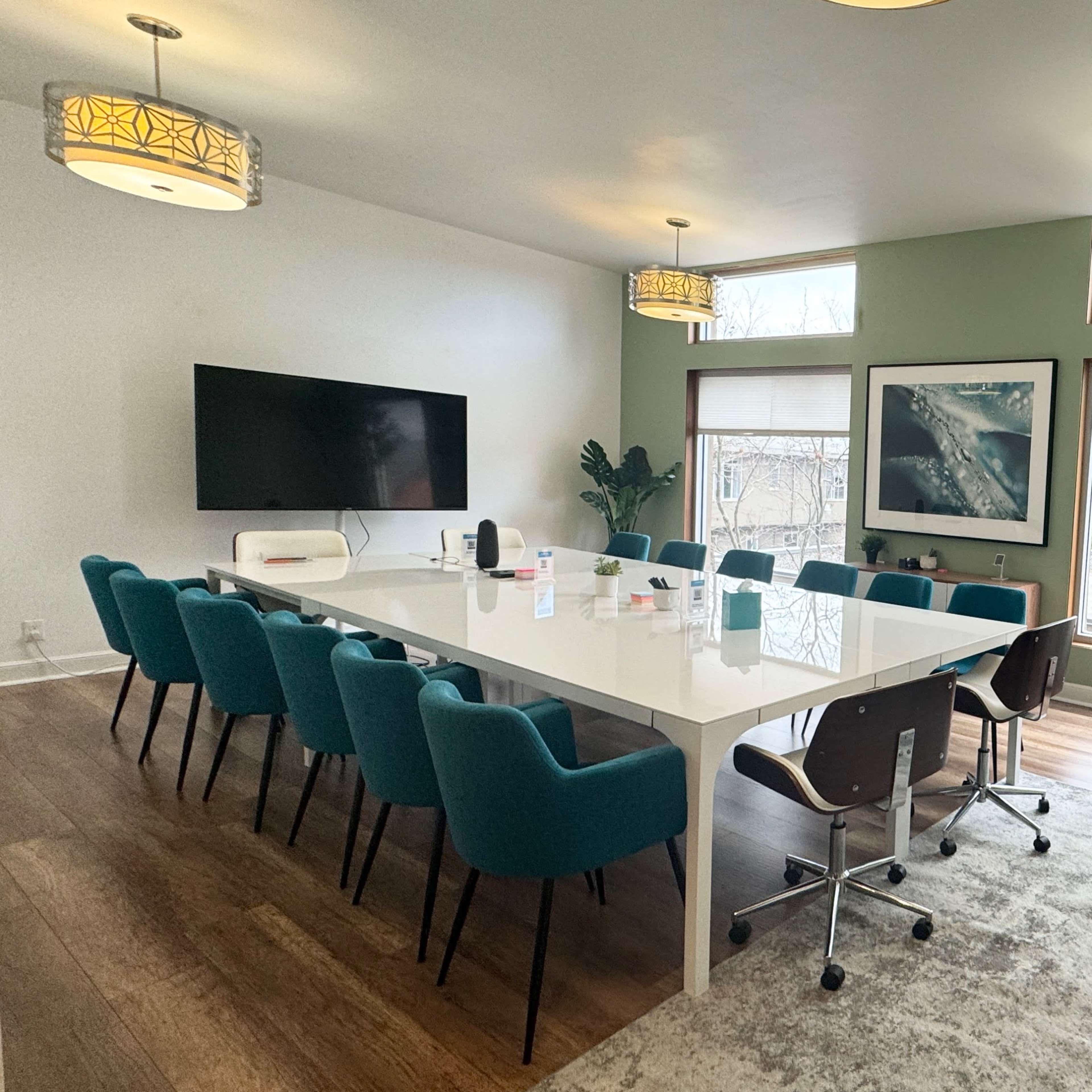 A modern conference room features a long white table surrounded by teal chairs, with large windows letting in natural light and greenery visible outside.
