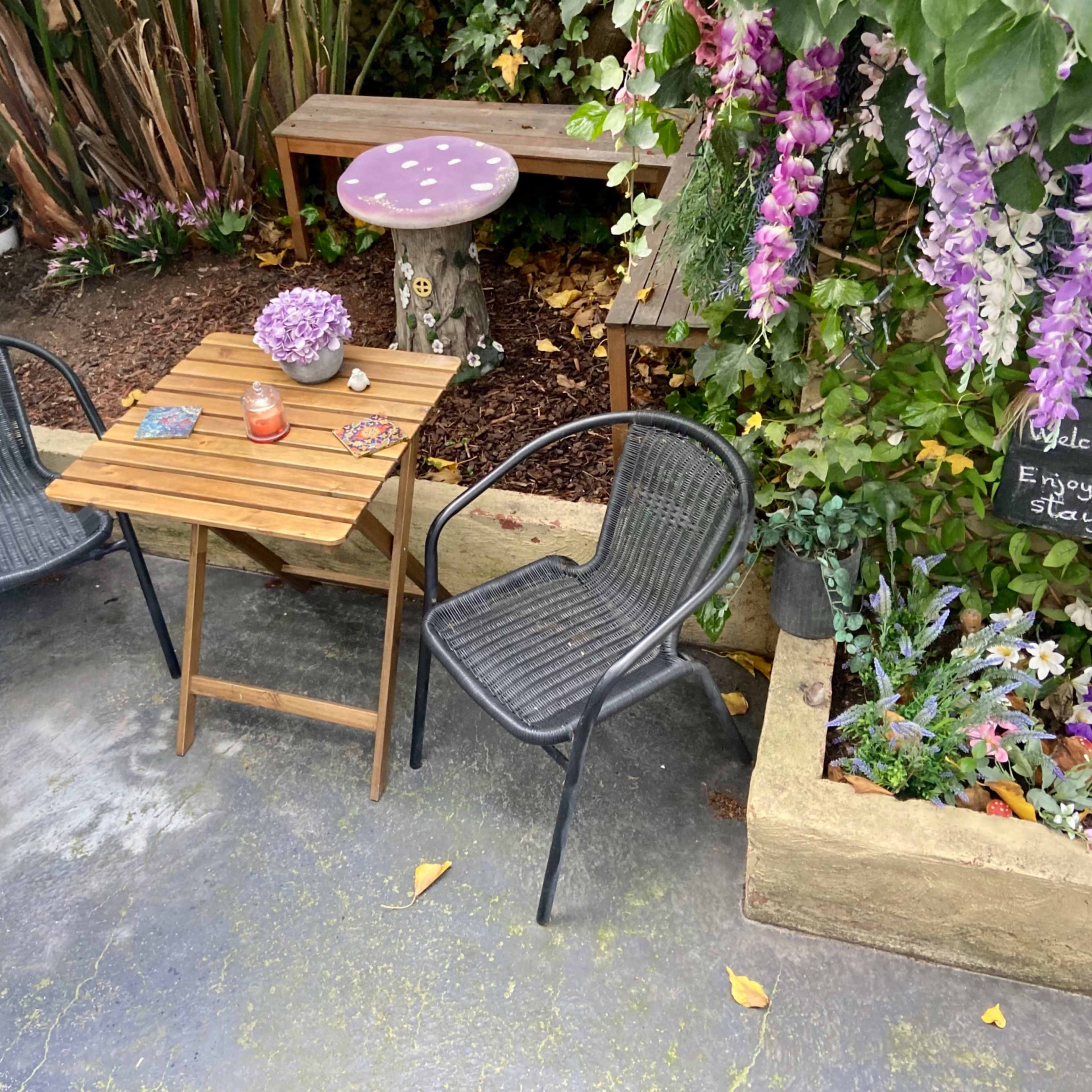 A small outdoor seating area features two black chairs, a wooden table, and a decorative mushroom stool surrounded by colorful flowers and greenery.