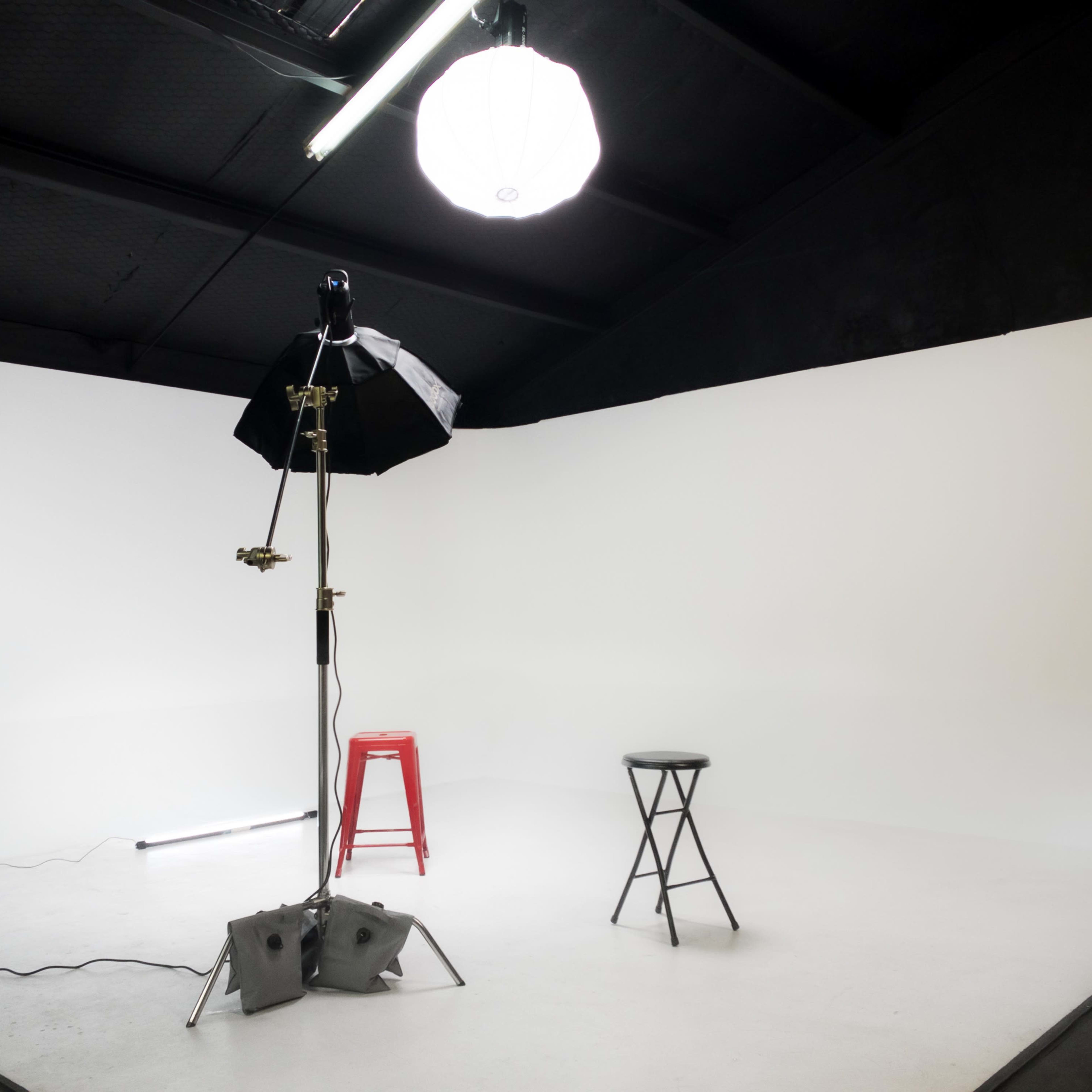 A minimalistic photography studio with two stools, a softbox light, and a spotlight on a white backdrop.