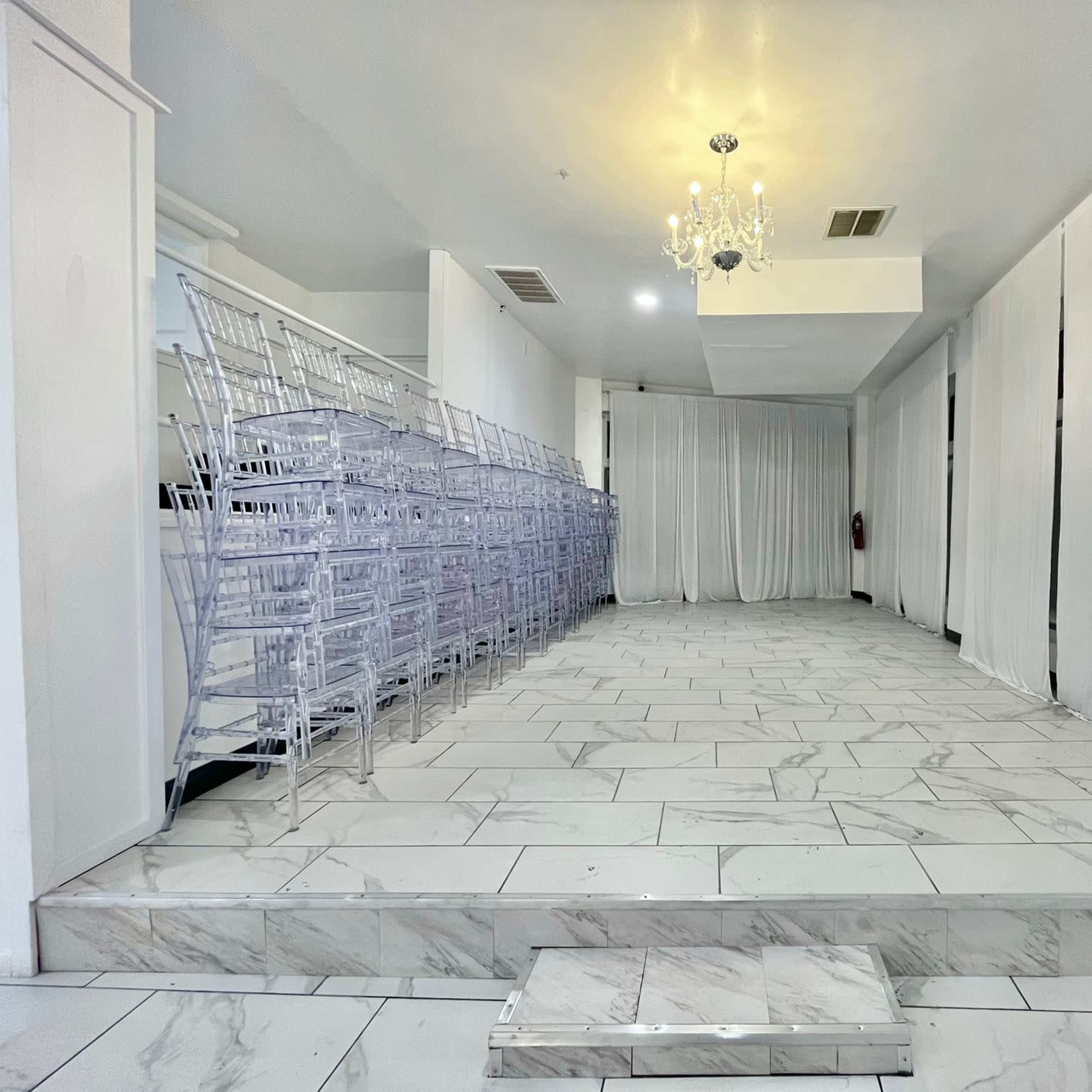 A spacious, well-lit room with white drapes, marble flooring, and stacked transparent Chiavari chairs along one side.