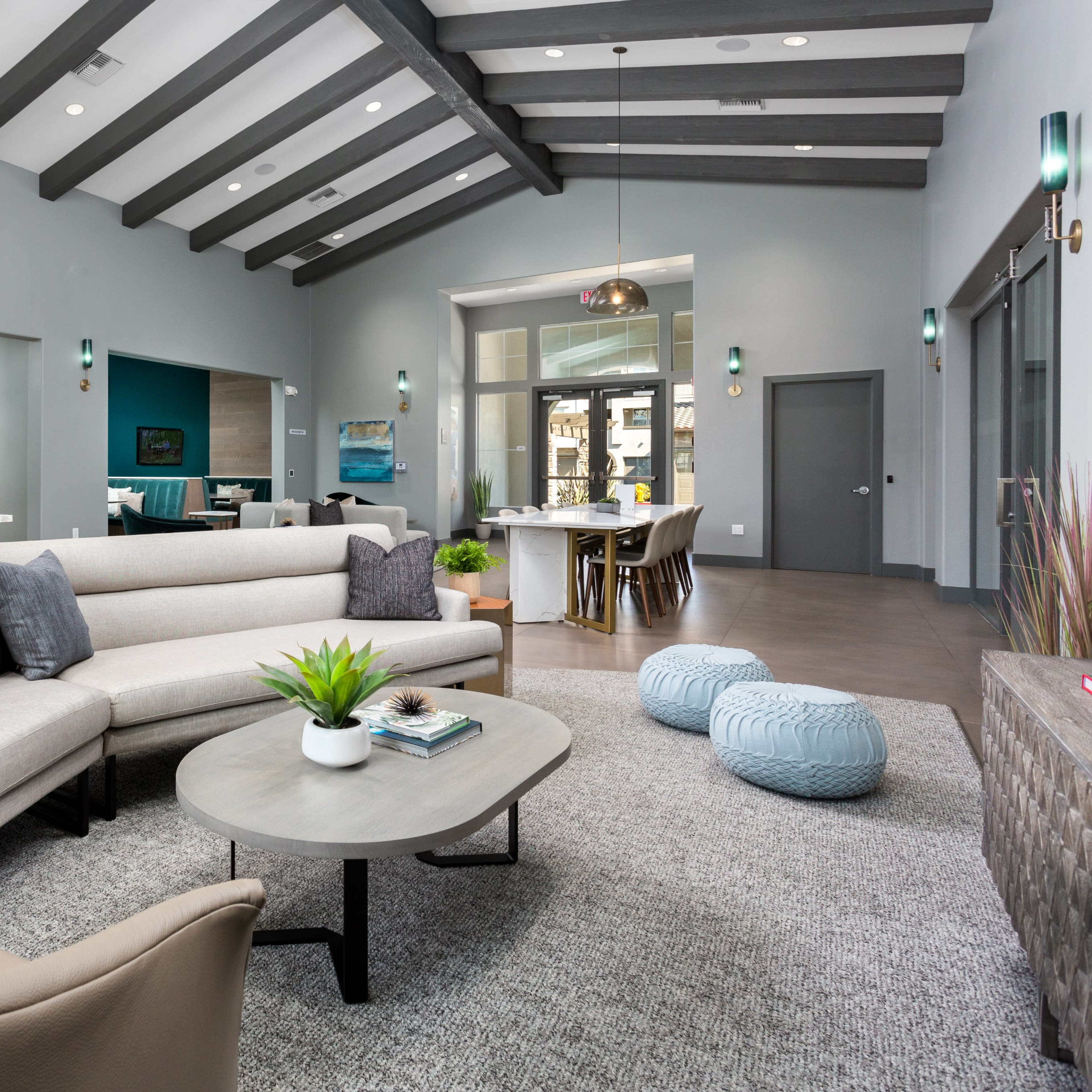 A modern living space features a sectional sofa, a coffee table, and a dining area with a wooden table and blue stools, surrounded by light gray walls and exposed wooden beams.