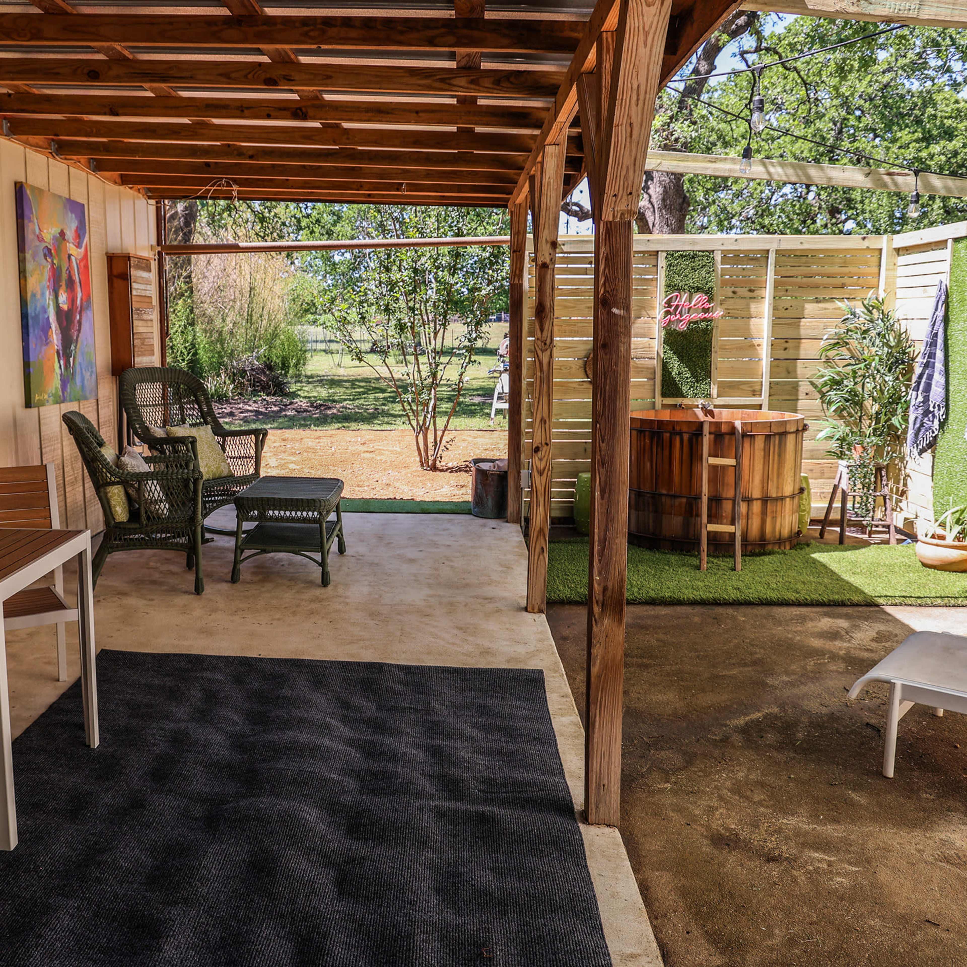 A covered outdoor space with a seating area, a small table, and a wooden hot tub in a landscaped yard.