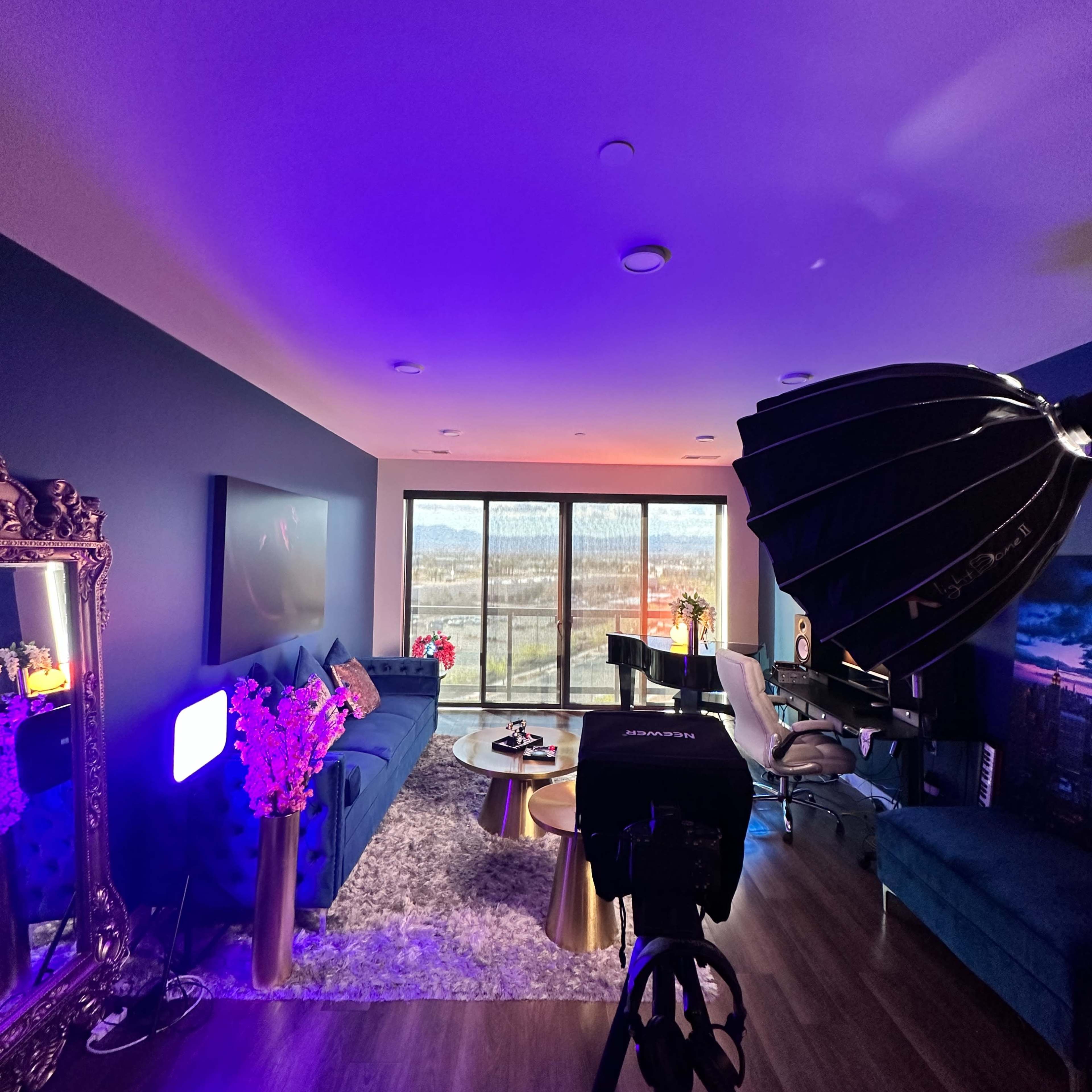 A modern living room features a blue couch, a grand piano, large windows with a view, and a camera setup with soft lighting.