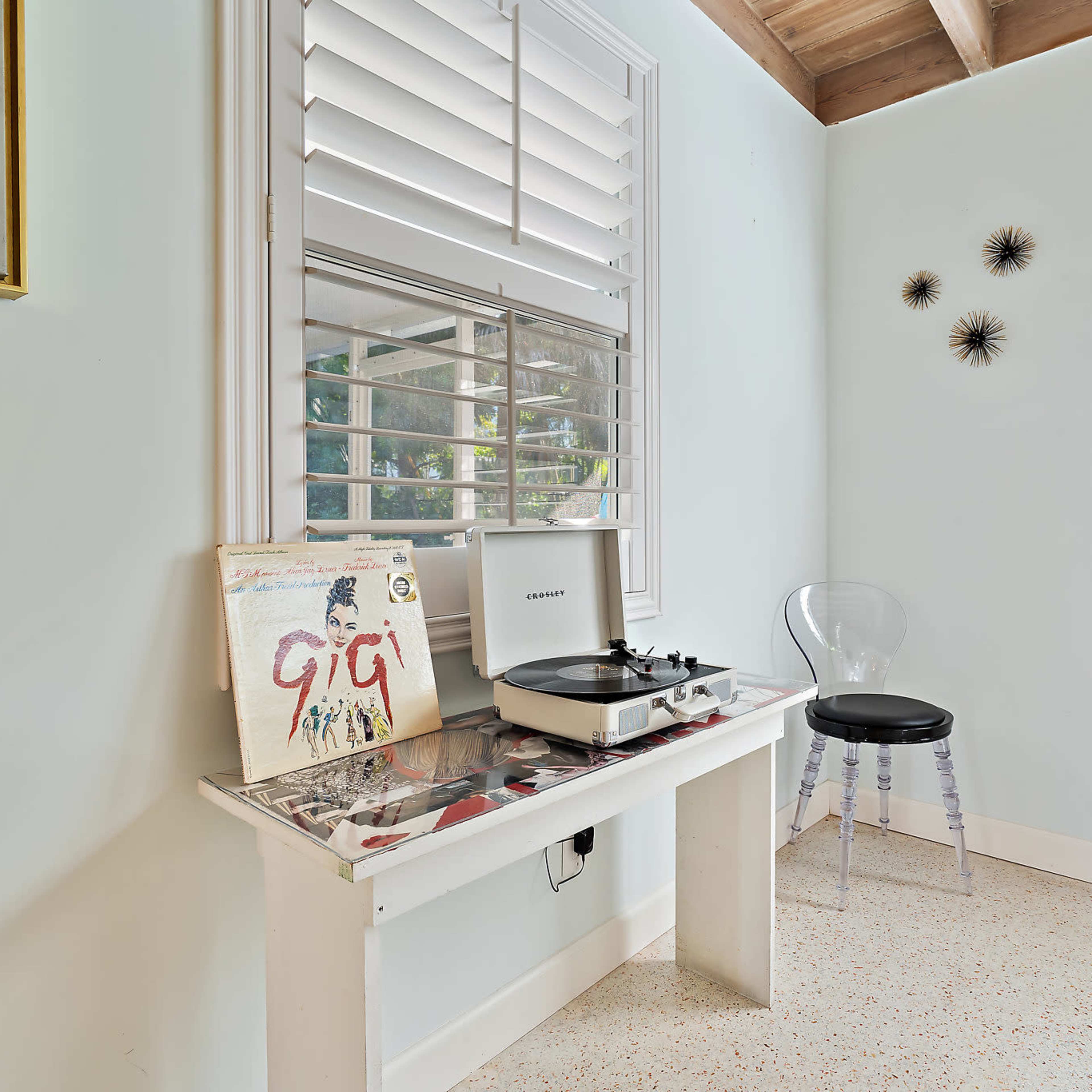 A record player and a vinyl album are placed on a white table beside a clear acrylic chair in a softly lit room with shuttered windows.