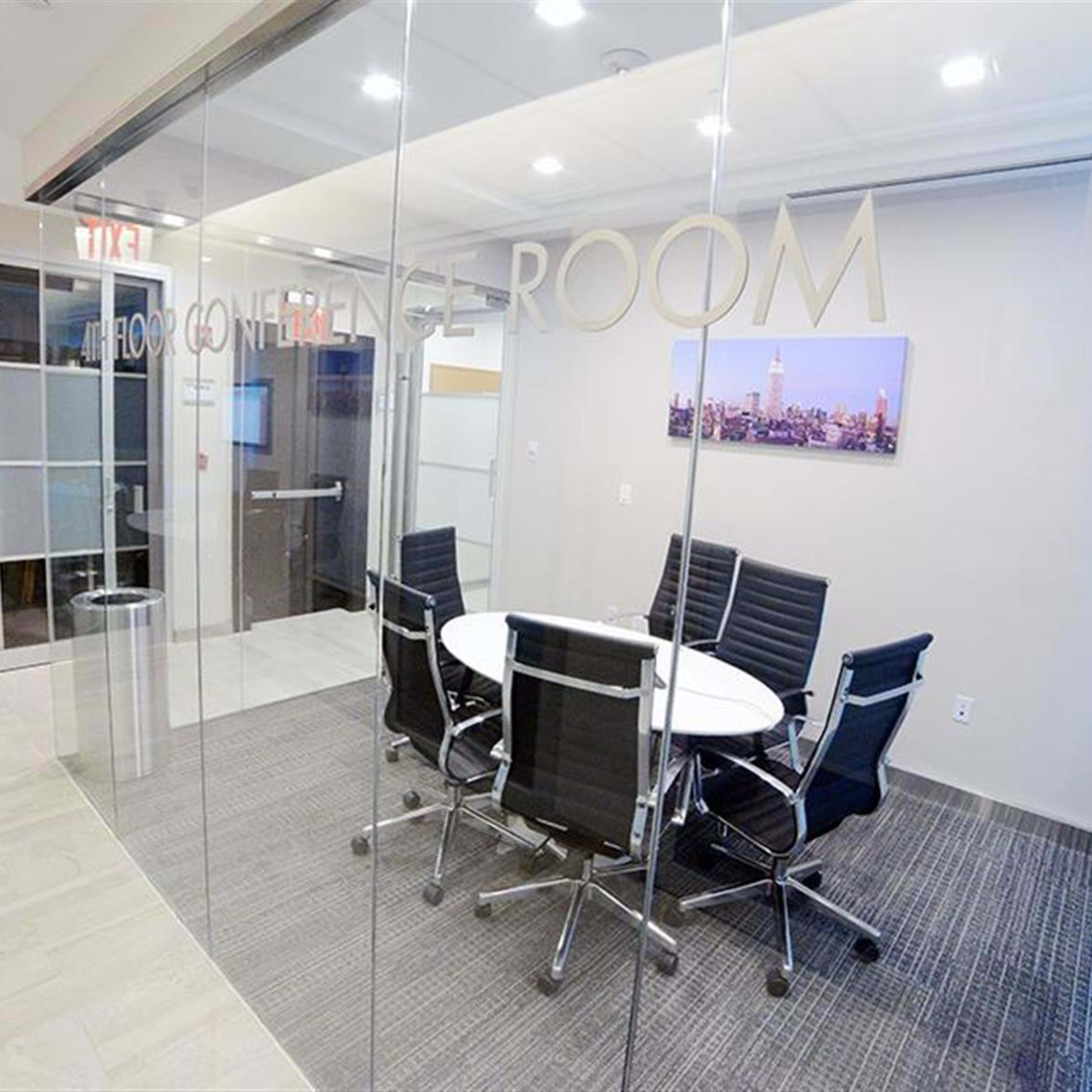 A modern conference room features a round table surrounded by black chairs, glass walls, and a large screen.