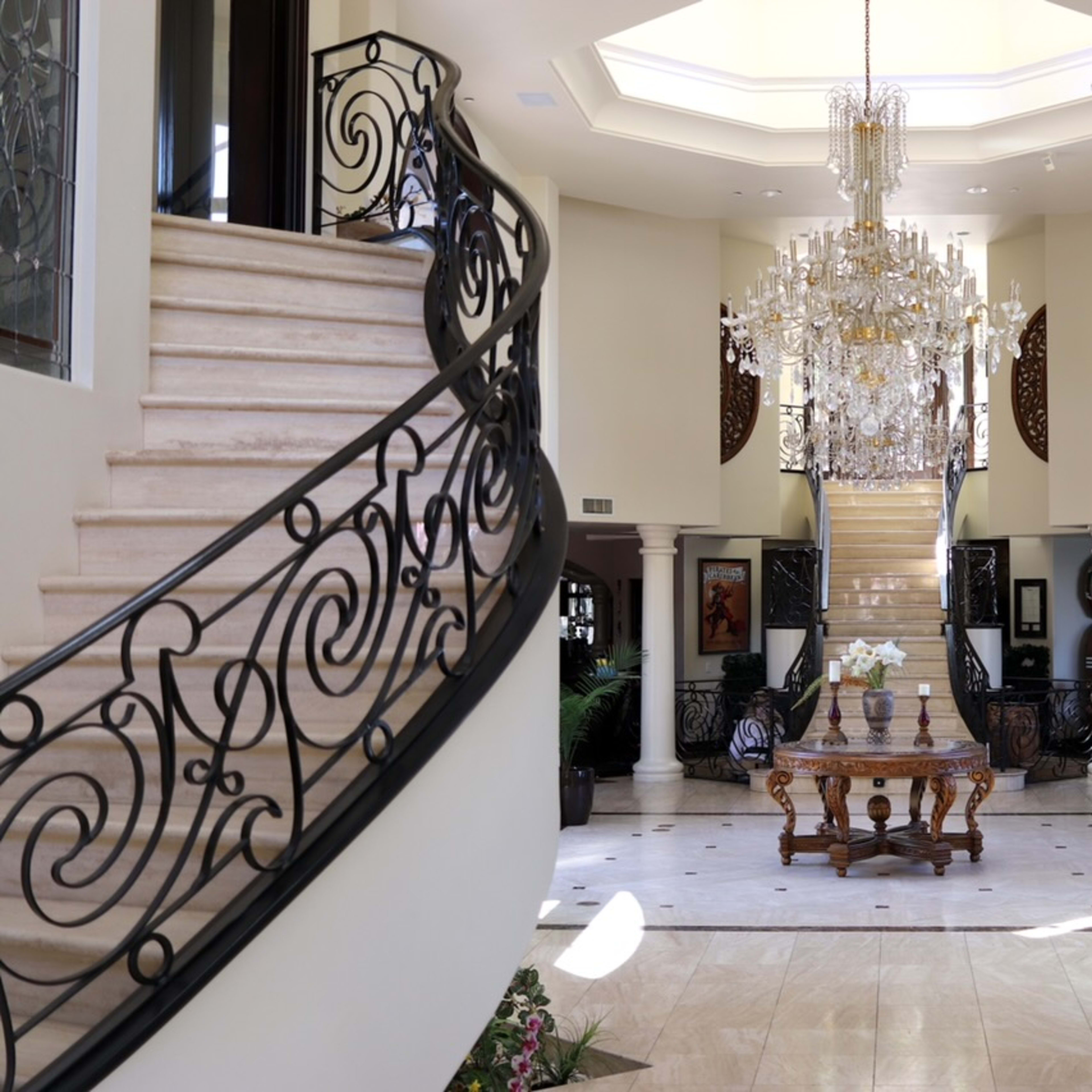 A grand foyer with a chandelier, a central wooden table, and a staircase, framed by curved staircases on either side.