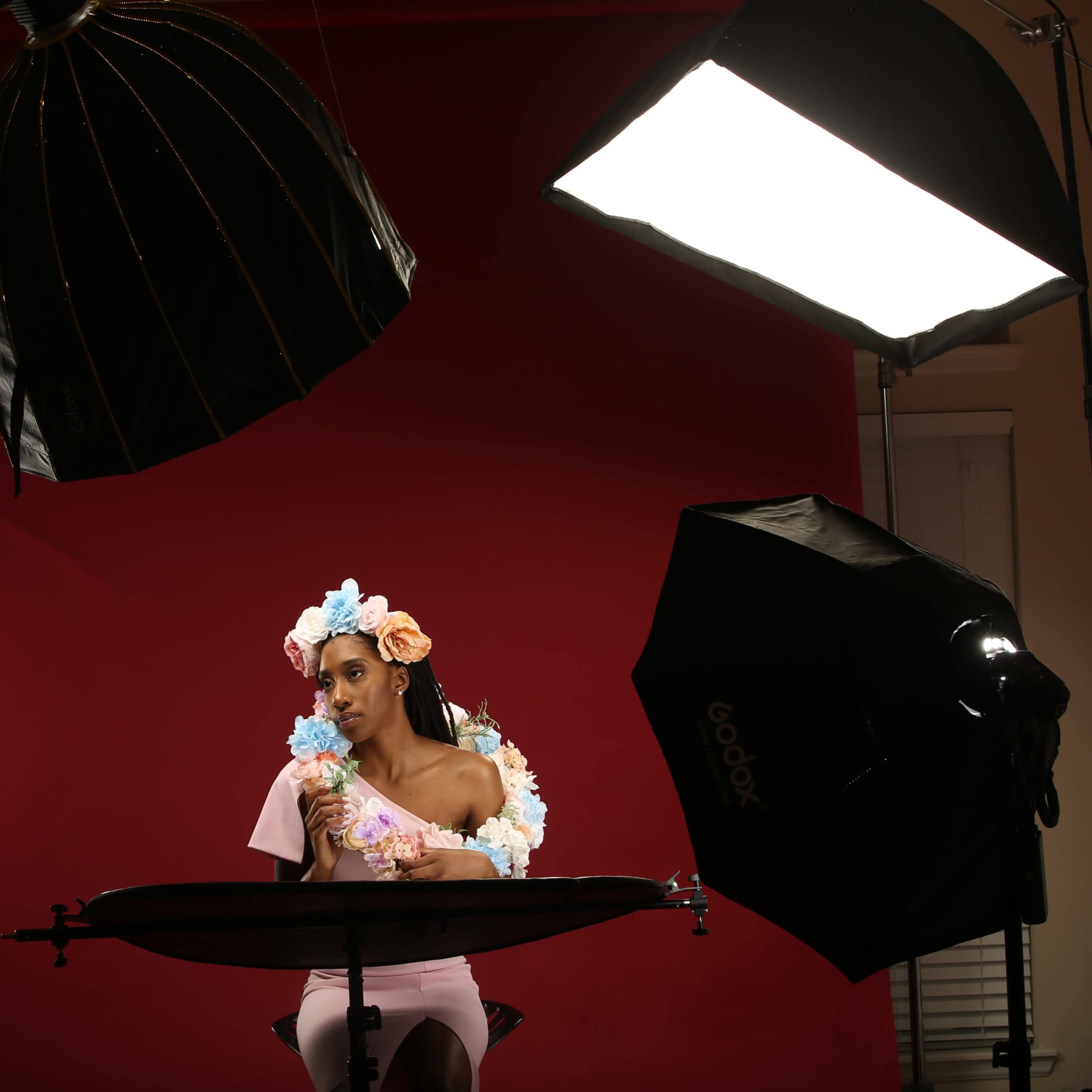 A model with a floral crown sits at a table in front of a red backdrop, surrounded by studio lights.
