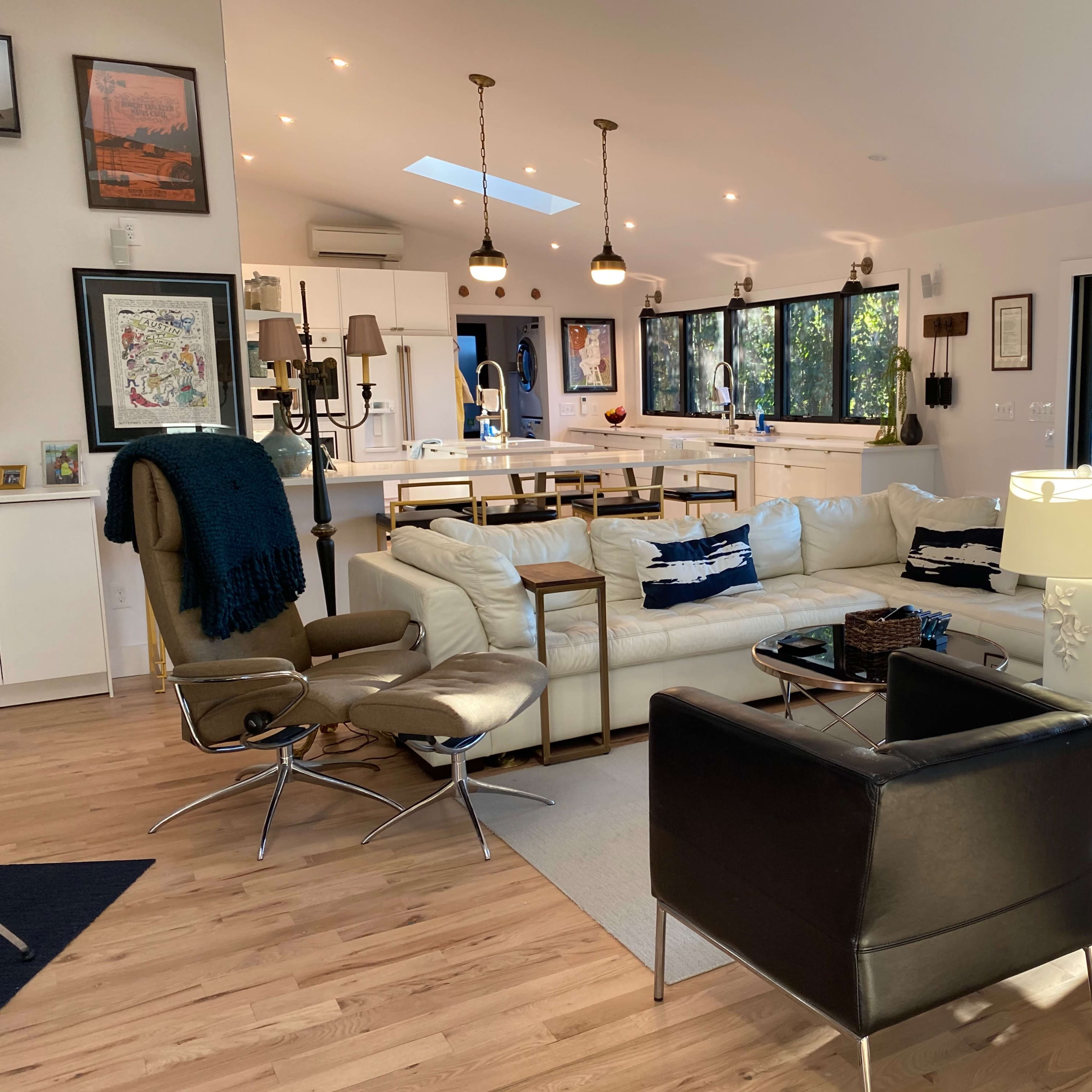 A modern, open-concept living room with a white sectional sofa, two black armchairs, and a wooden dining area in the background.