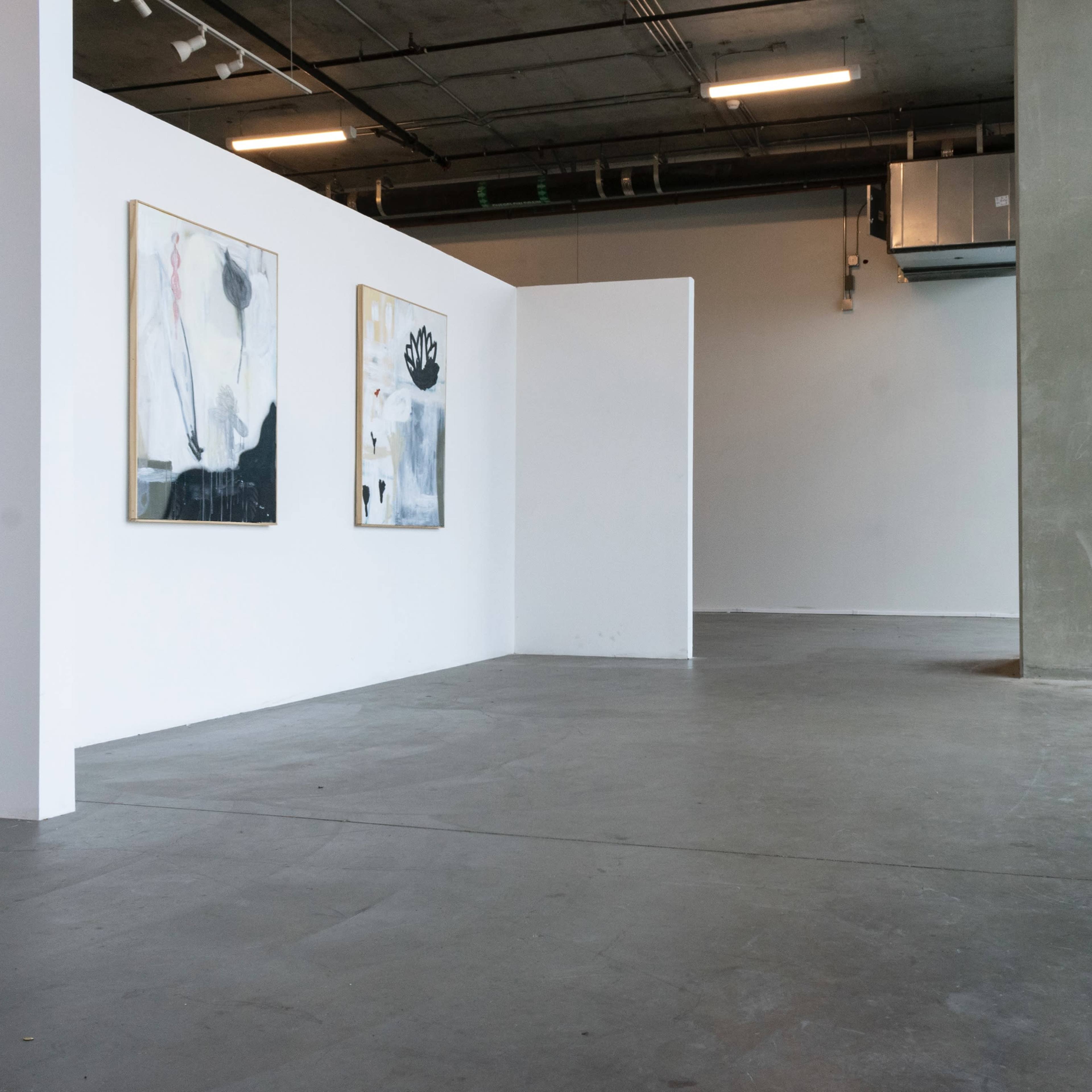 A spacious interior of an art gallery featuring two abstract paintings on a white wall and large windows allowing natural light to enter.