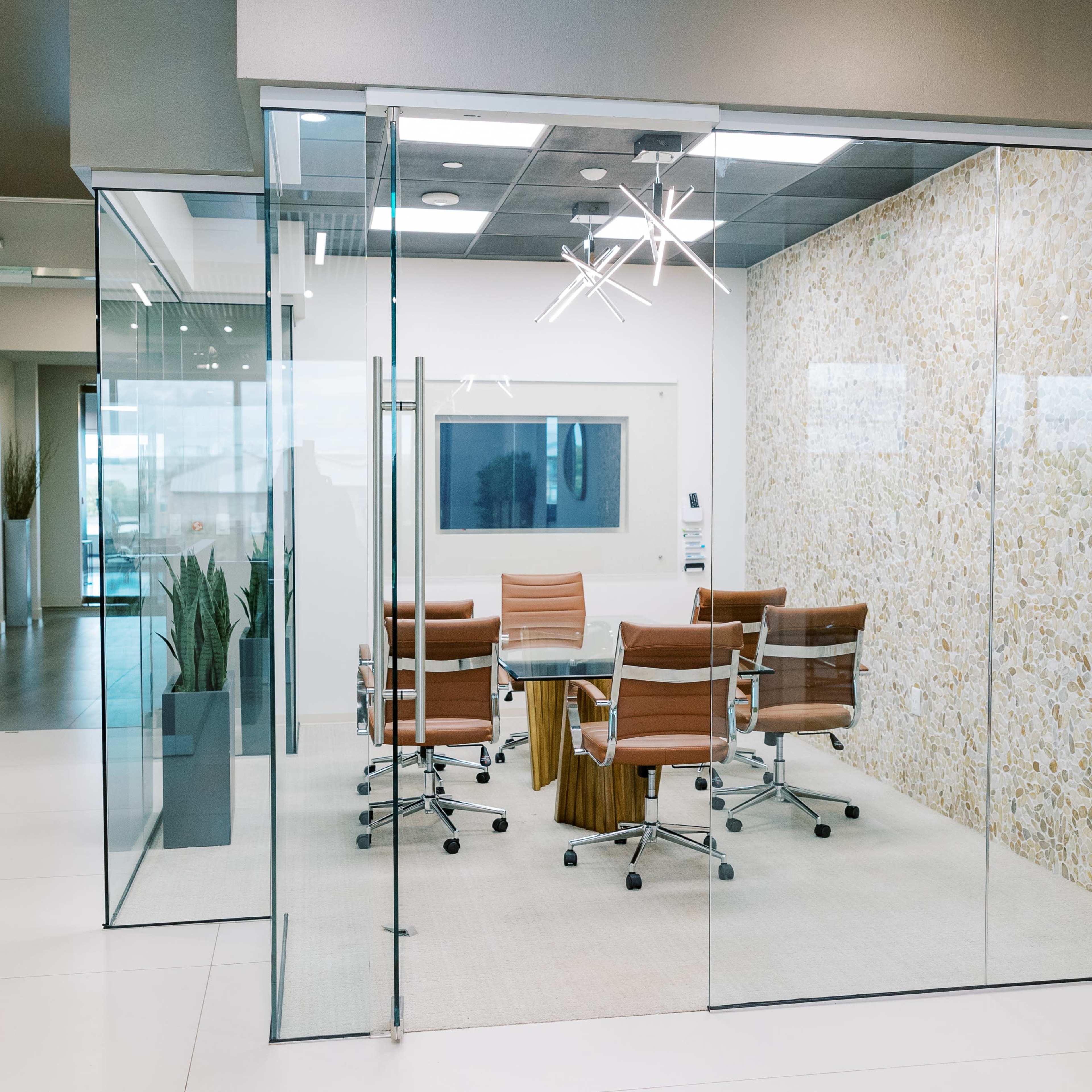 A glass-walled conference room contains a round table surrounded by four brown leather chairs, with a large light fixture hanging above.