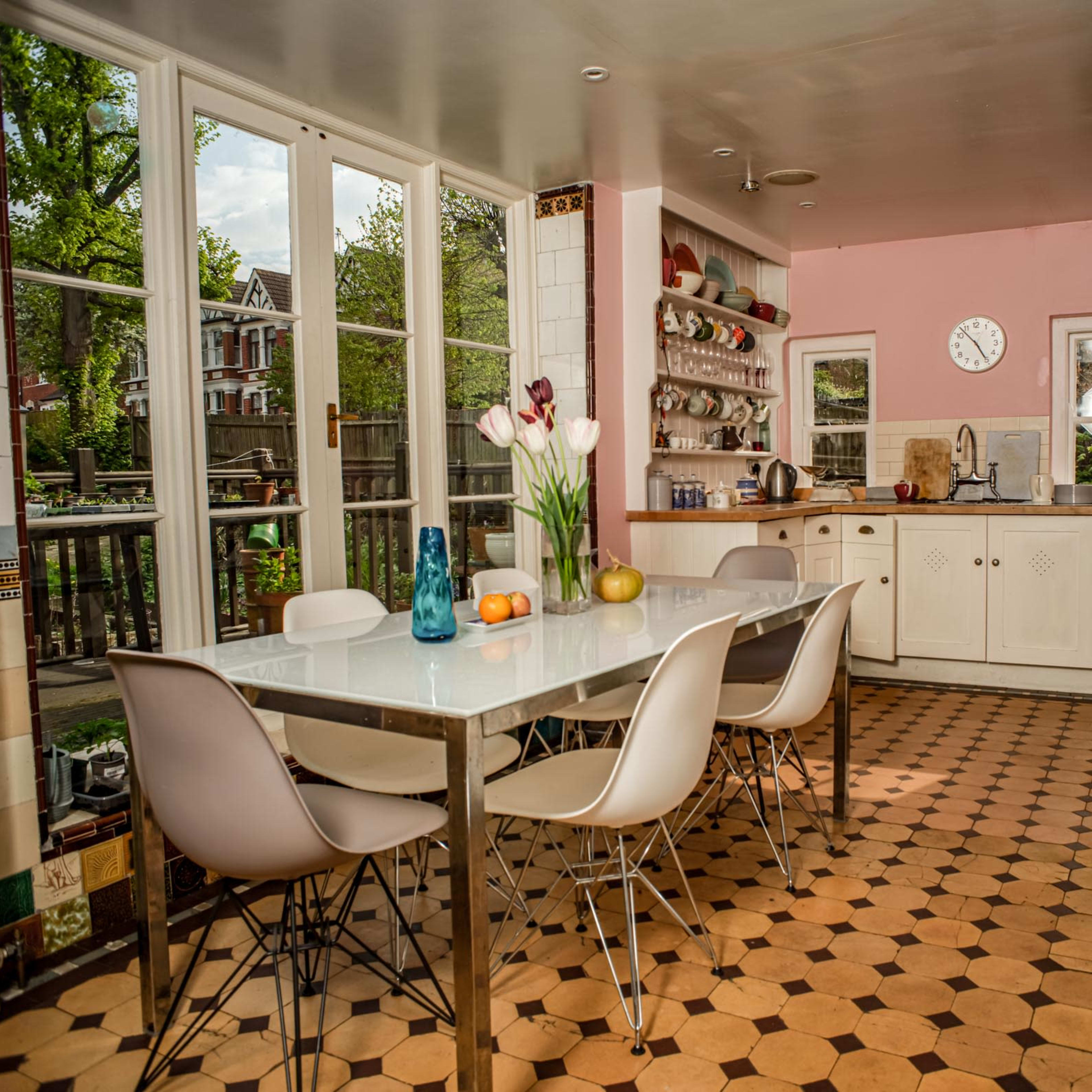 A bright kitchen features a large table surrounded by modern chairs, with dishes and a vase of flowers on the table, and windows letting in natural light.