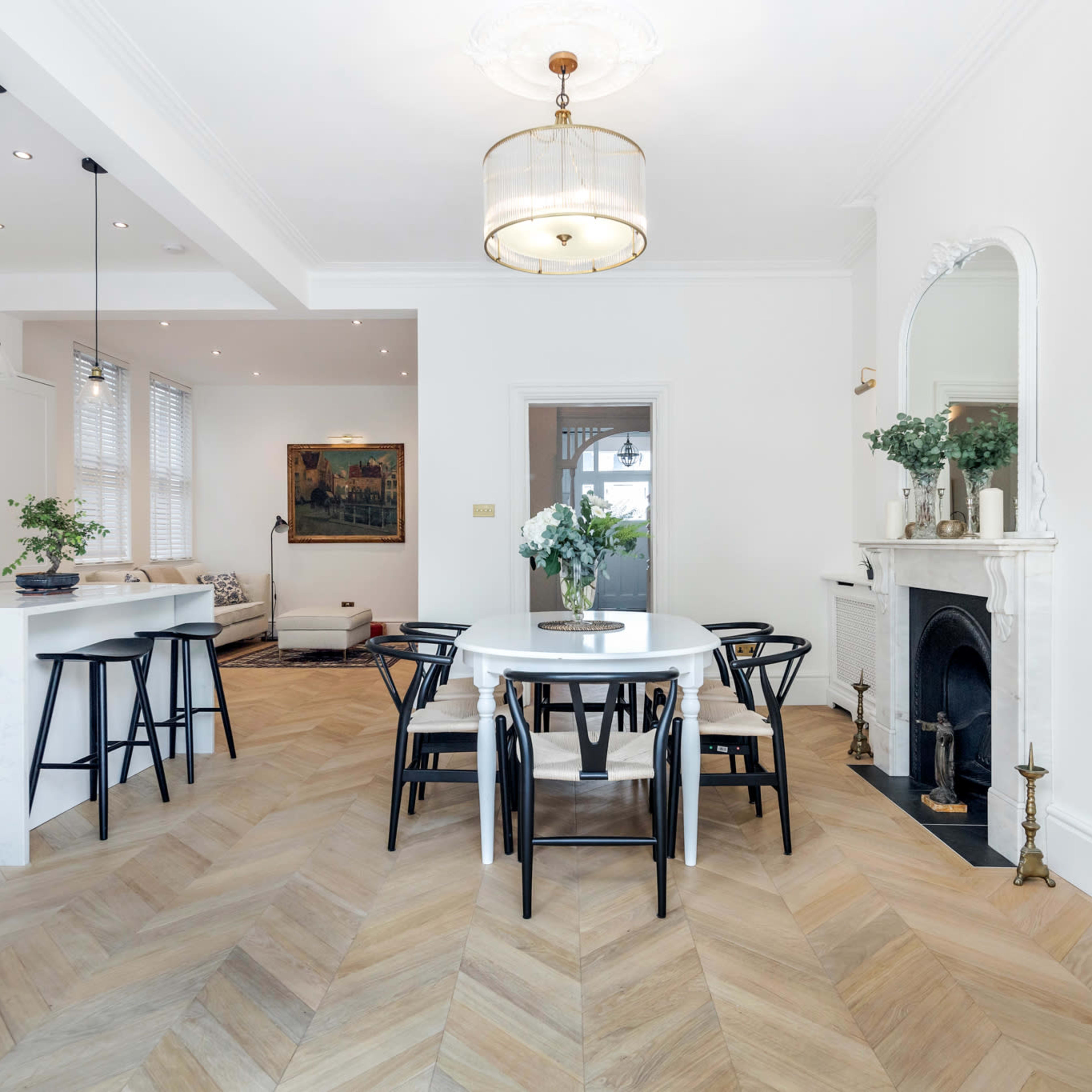 A modern dining area with a circular table, black chairs, and a light fixture, featuring herringbone-patterned hardwood flooring and a fireplace with decorative elements.