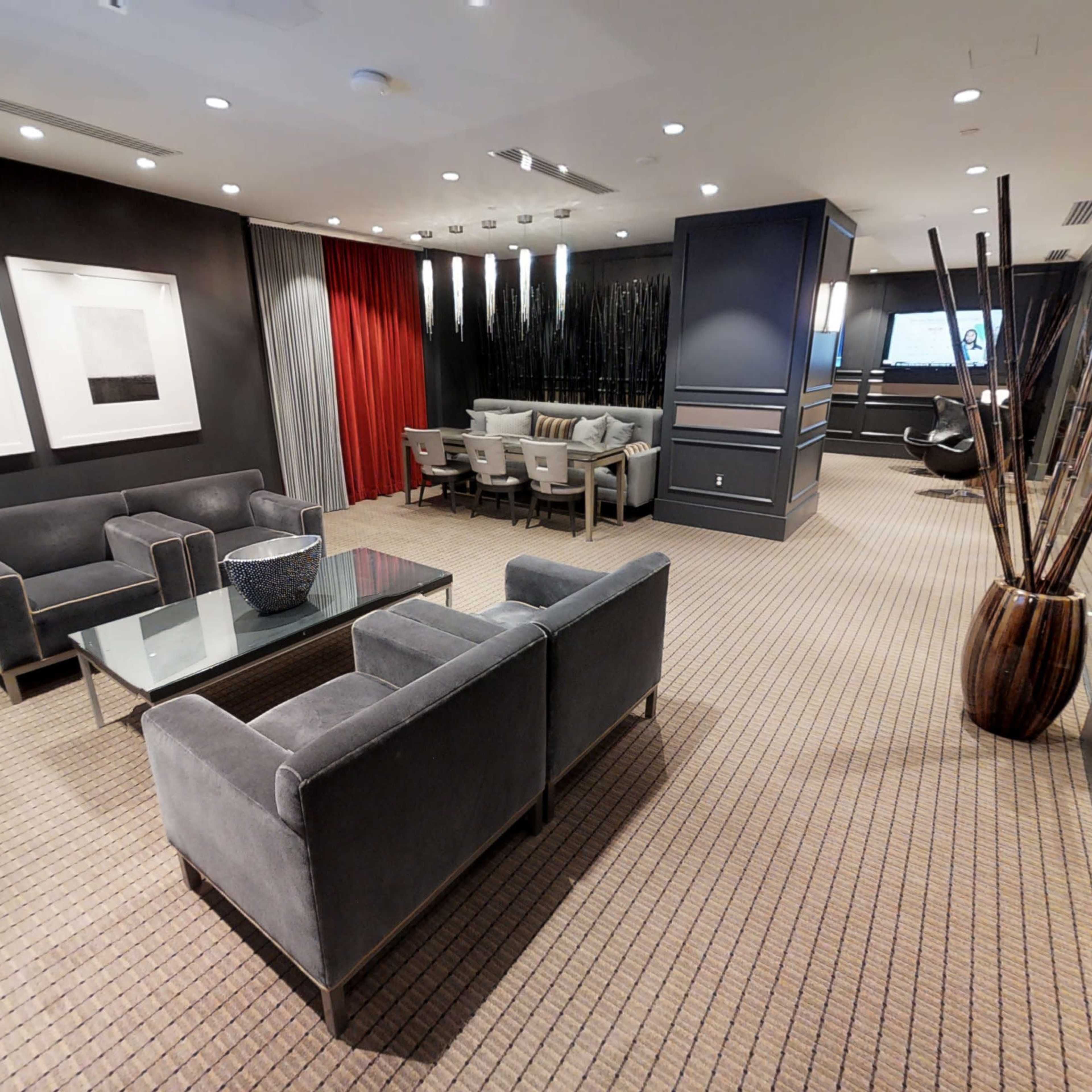 A modern lounge area with gray sofas, a glass coffee table, a dining table with chairs, and decorative plants against a backdrop of dark walls and a contrasting red curtain.