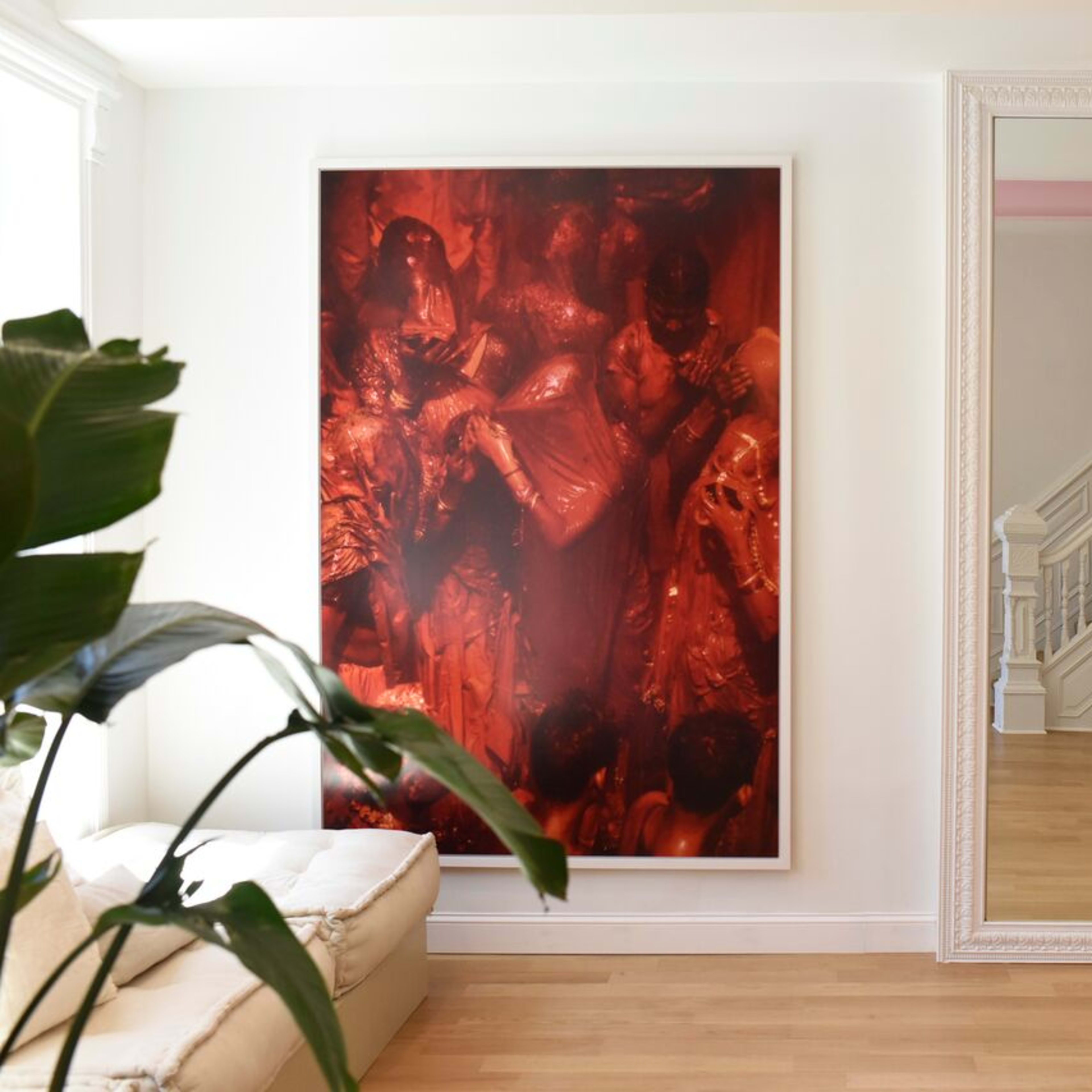 A bright interior with a large red-toned artwork on the wall, a mirror reflecting a staircase, and a decorative candelabra beside a light-colored sofa.