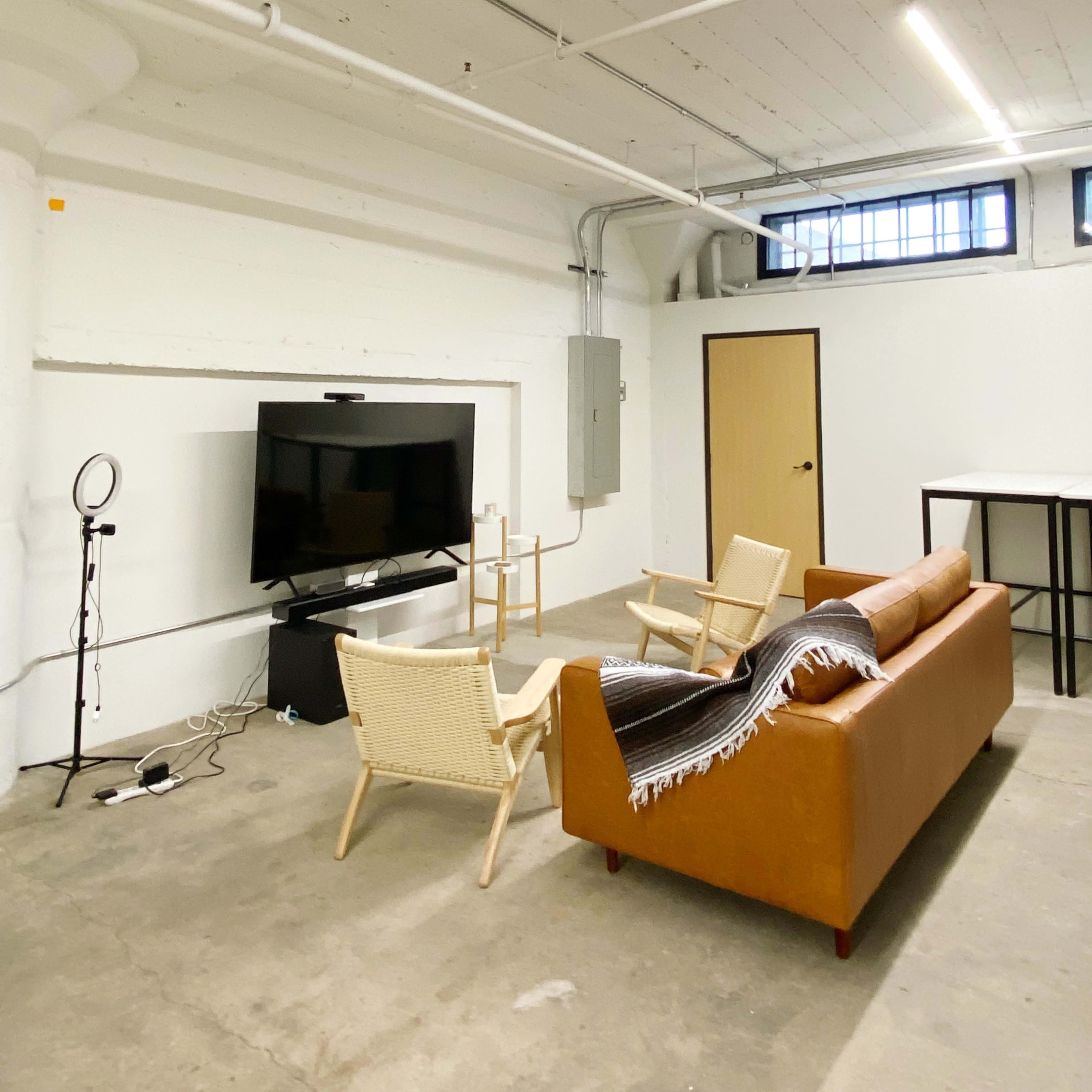 A modern, minimalistic room with a brown leather sofa, two light-colored chairs, a large television on a stand, and a table against a white wall.