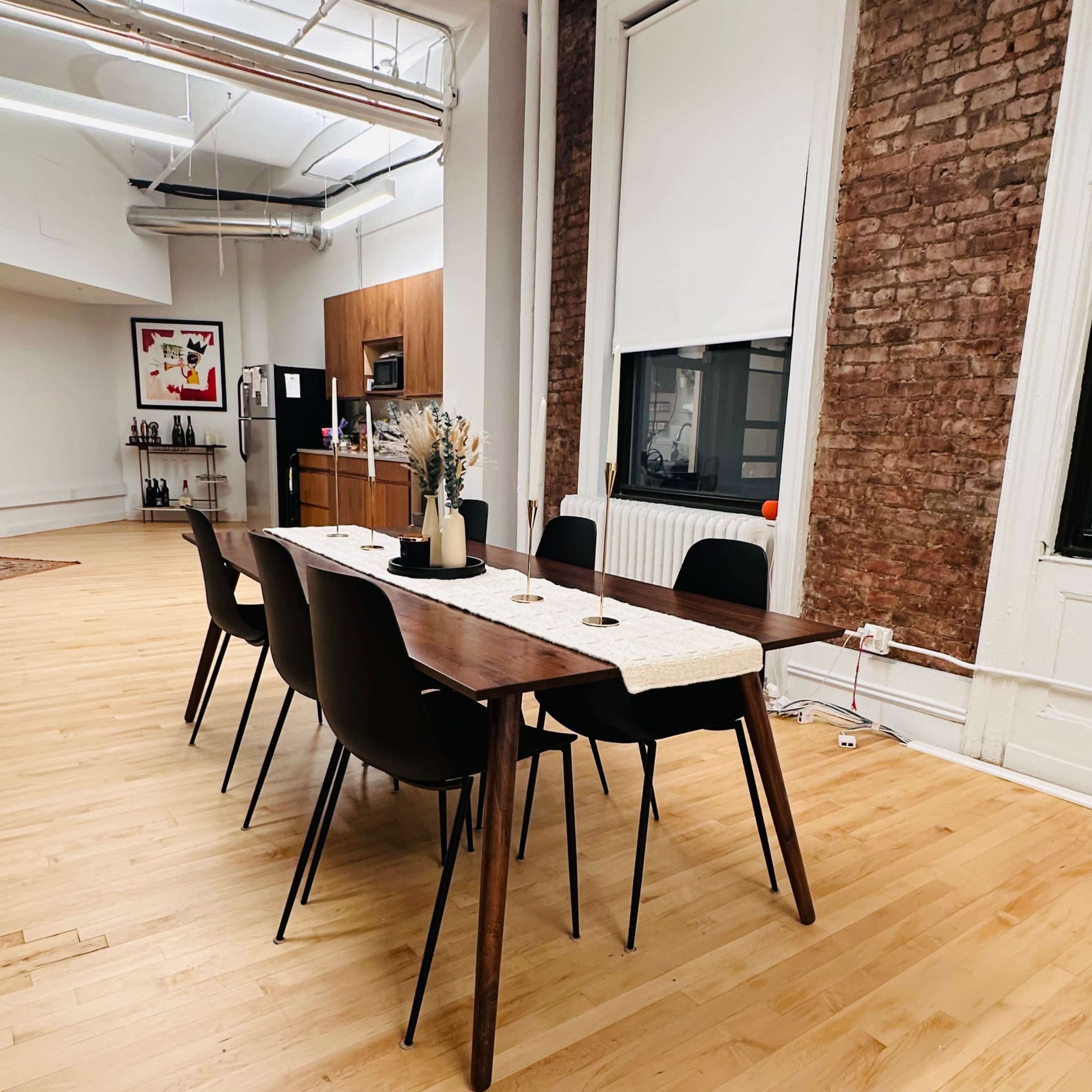 A long wooden dining table with six chairs is set in a spacious room featuring exposed brick walls and a kitchen area in the background.