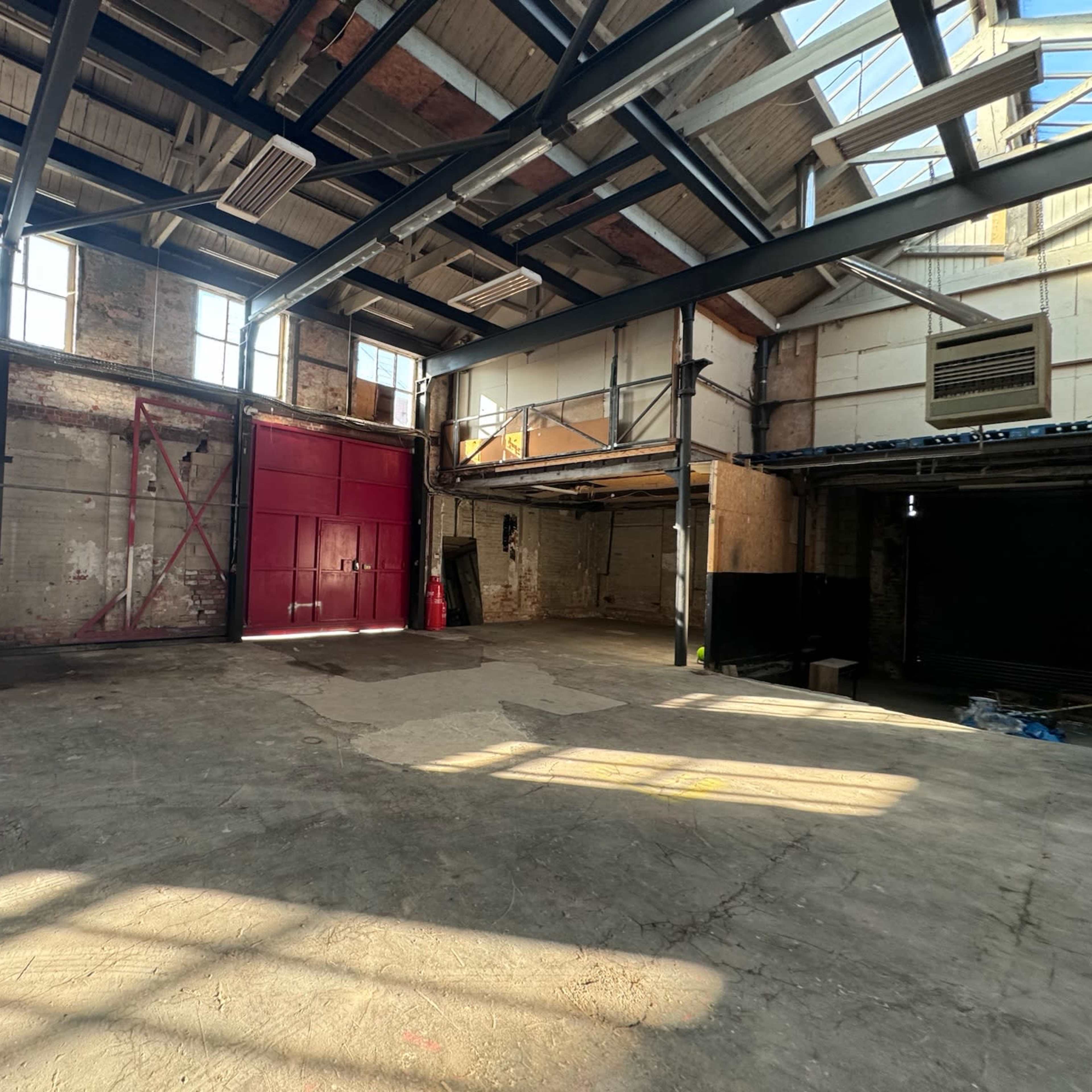 An empty industrial warehouse with high ceilings, exposed brick walls, and large windows allowing natural light to stream in.