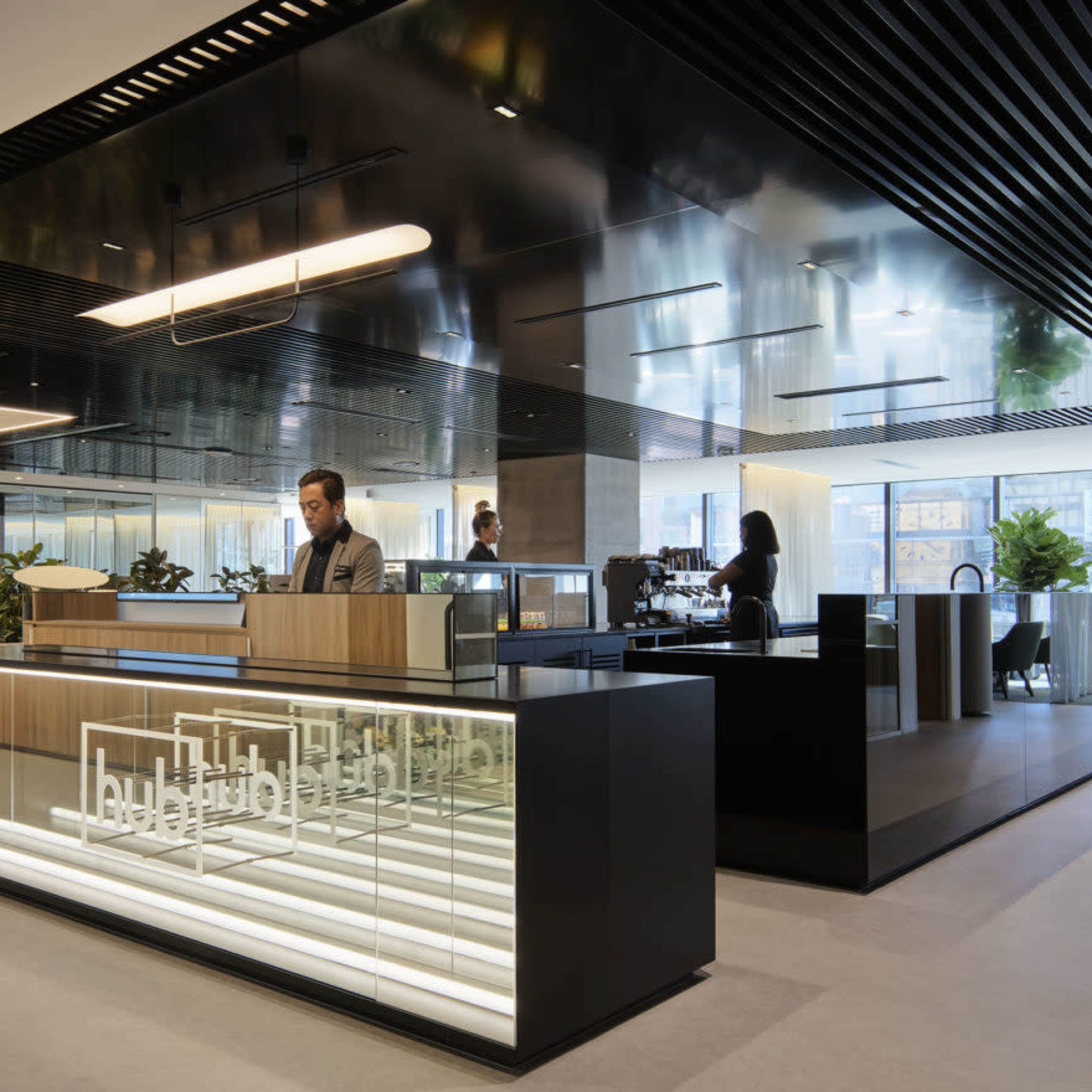 A modern office lobby with a reception area featuring a sleek desk and a staircase leading to an upper level, surrounded by greenery and large windows.