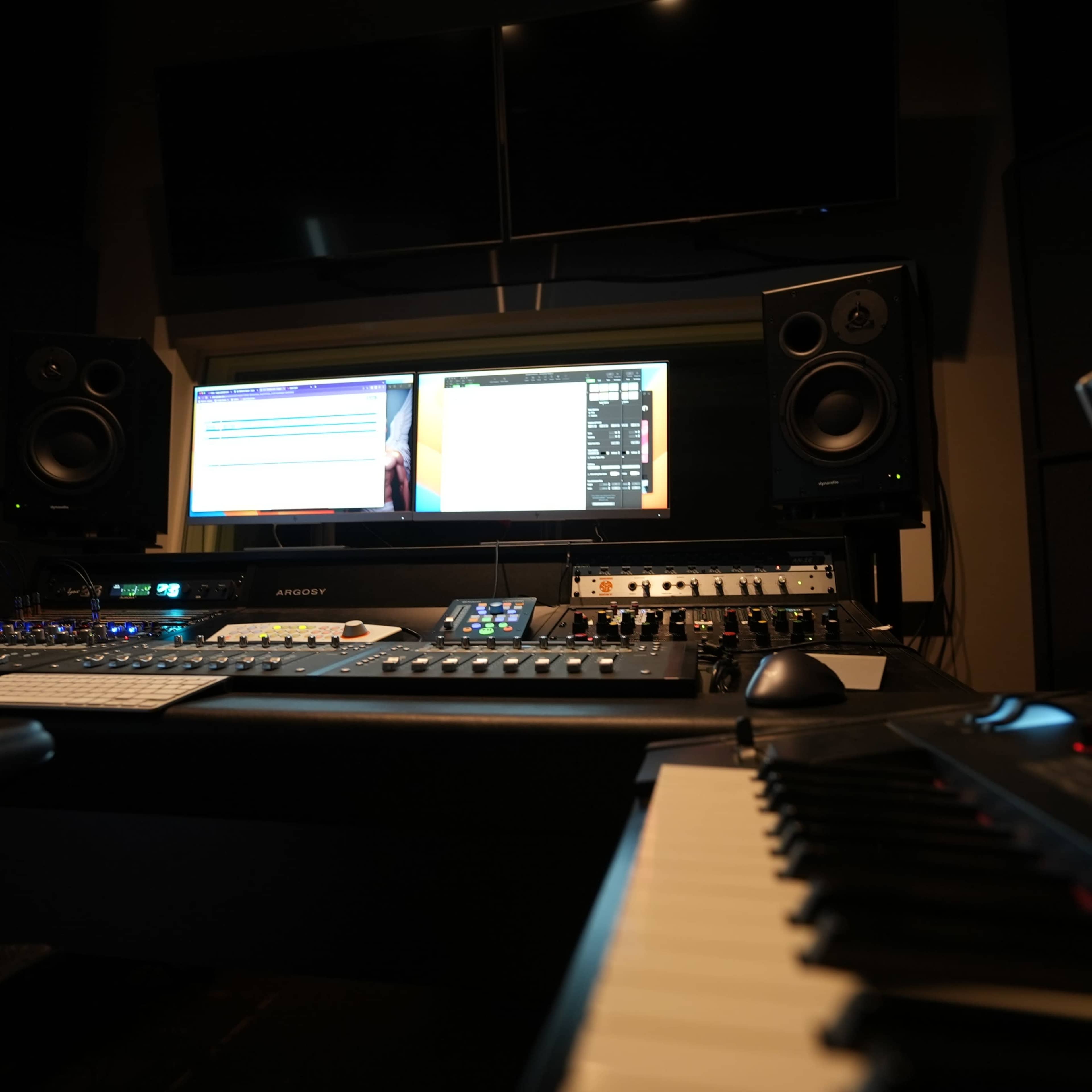 A music recording studio with a control desk featuring computer monitors, audio interfaces, and a keyboard.