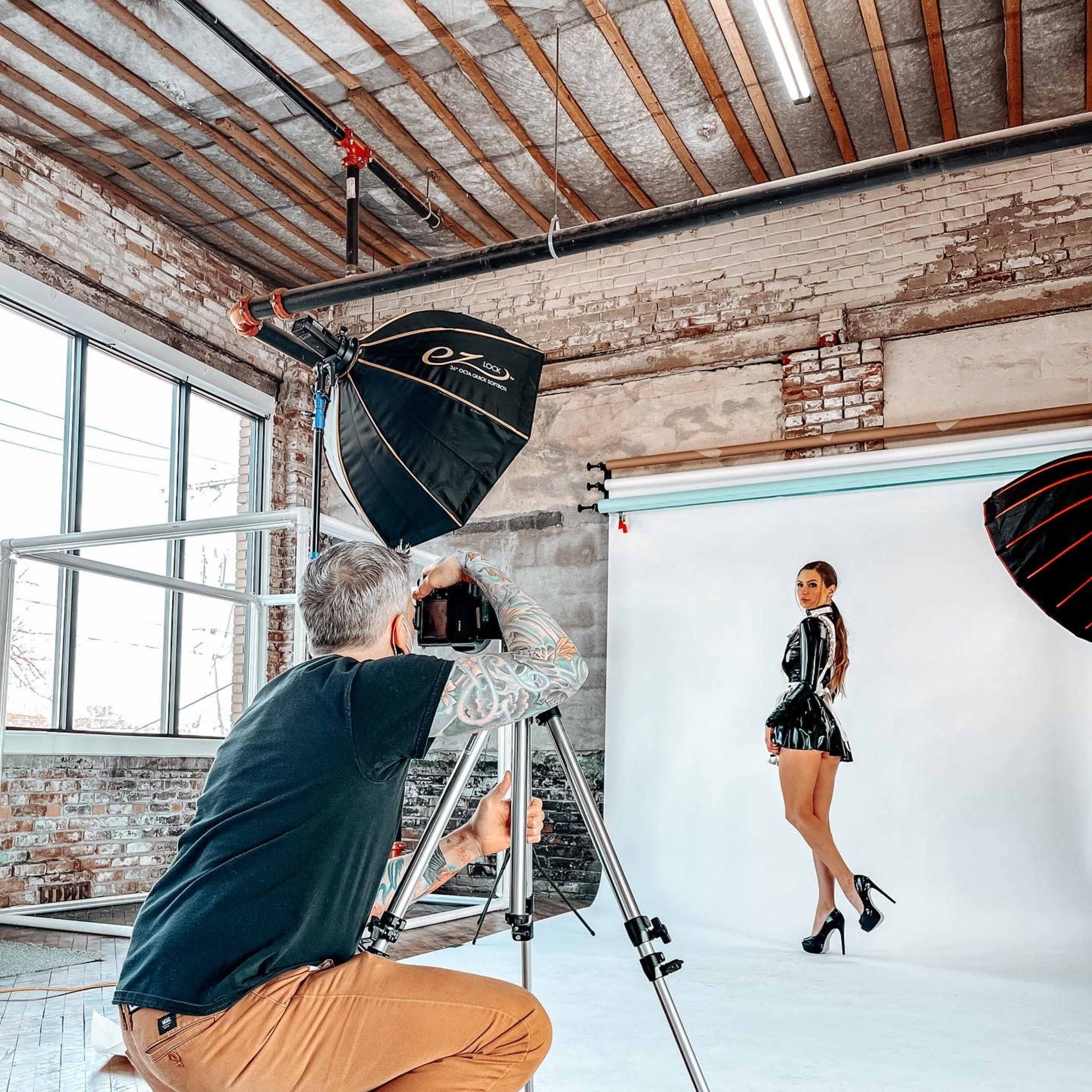 A photographer crouches behind a camera on a tripod, capturing a model posing in a shiny outfit against a light backdrop in a studio with exposed brick walls.