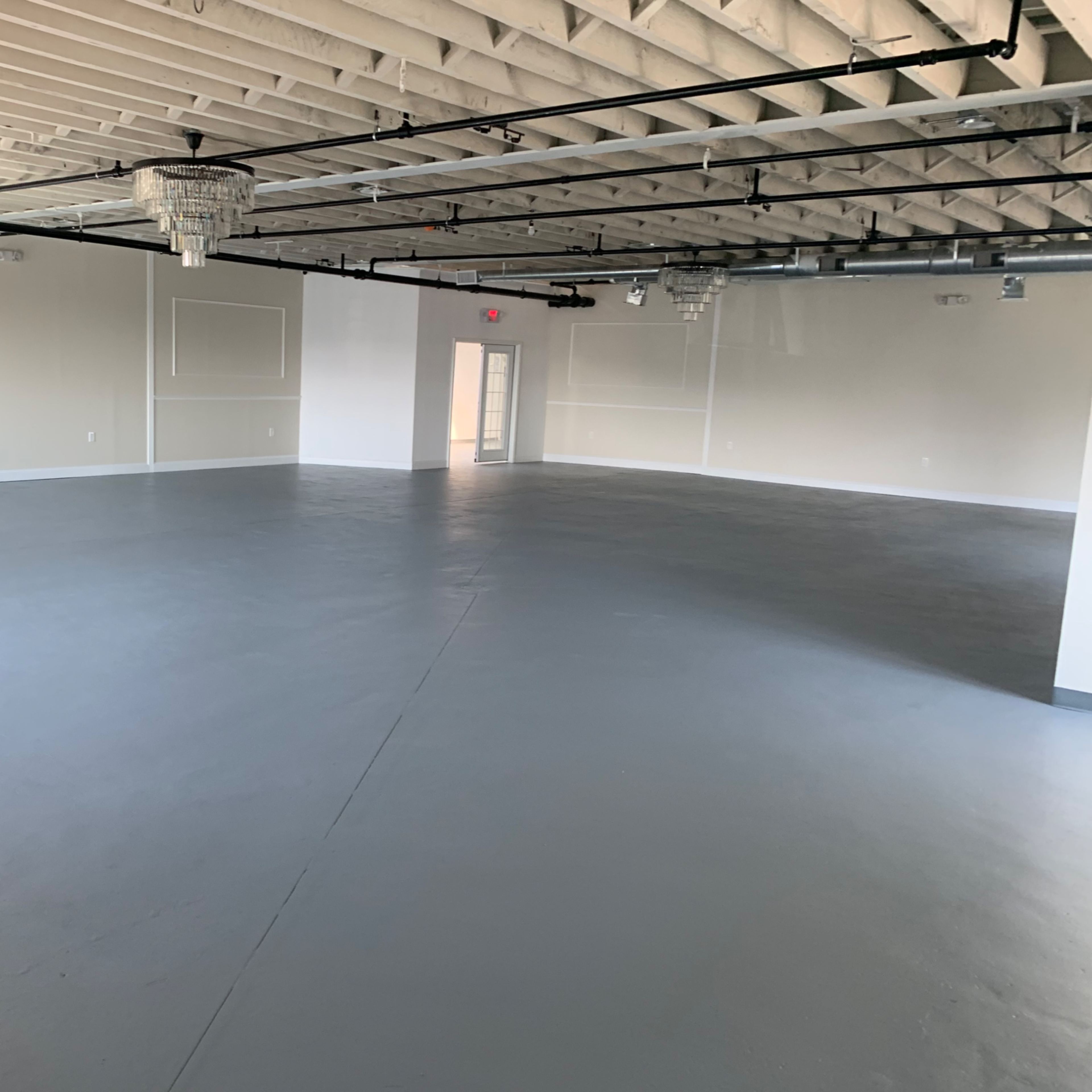 A large, empty room with a gray concrete floor and exposed ceiling beams, featuring several light fixtures and a door at one end.