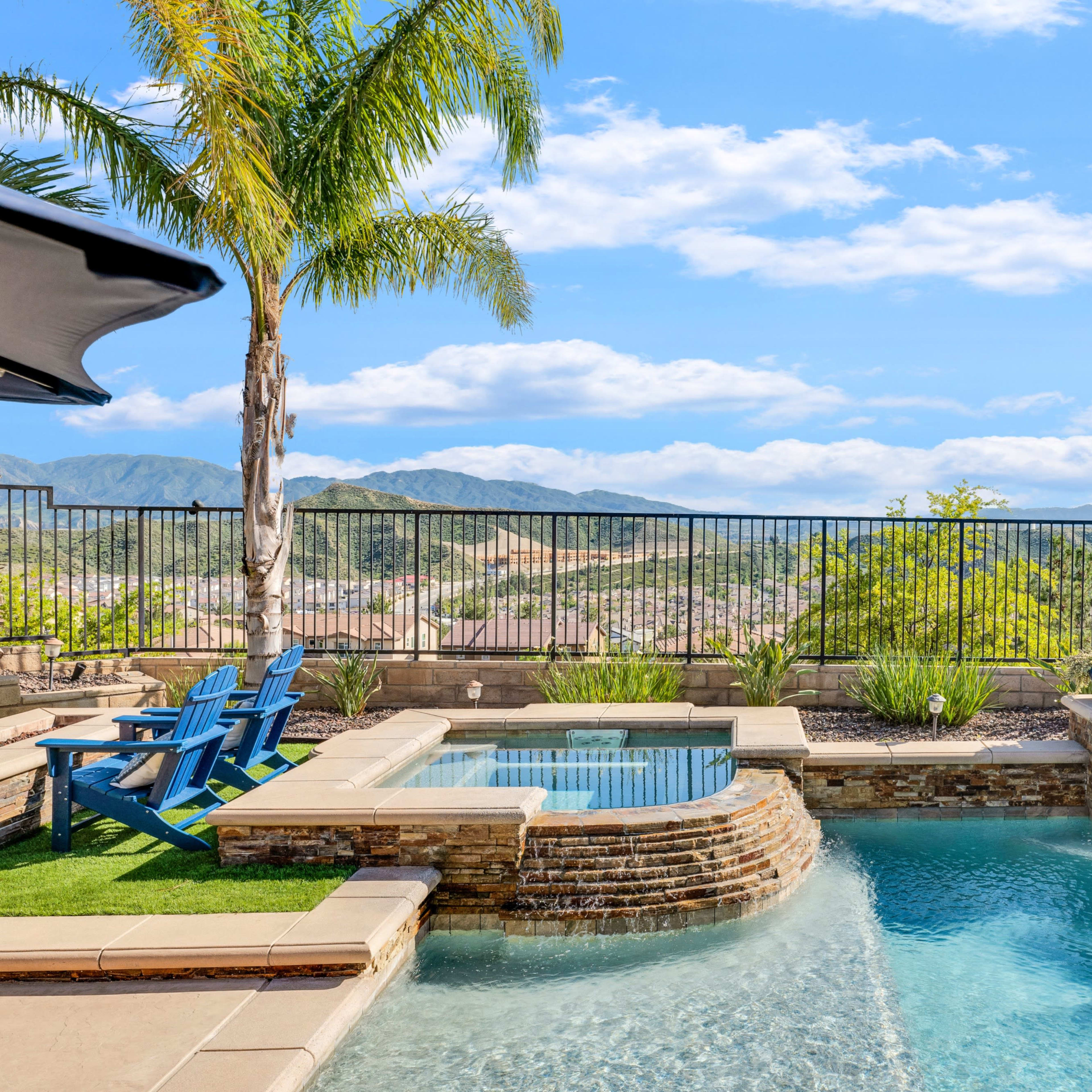A landscaped pool area with a spa, blue lounge chairs, and a panoramic view of mountains and sky.