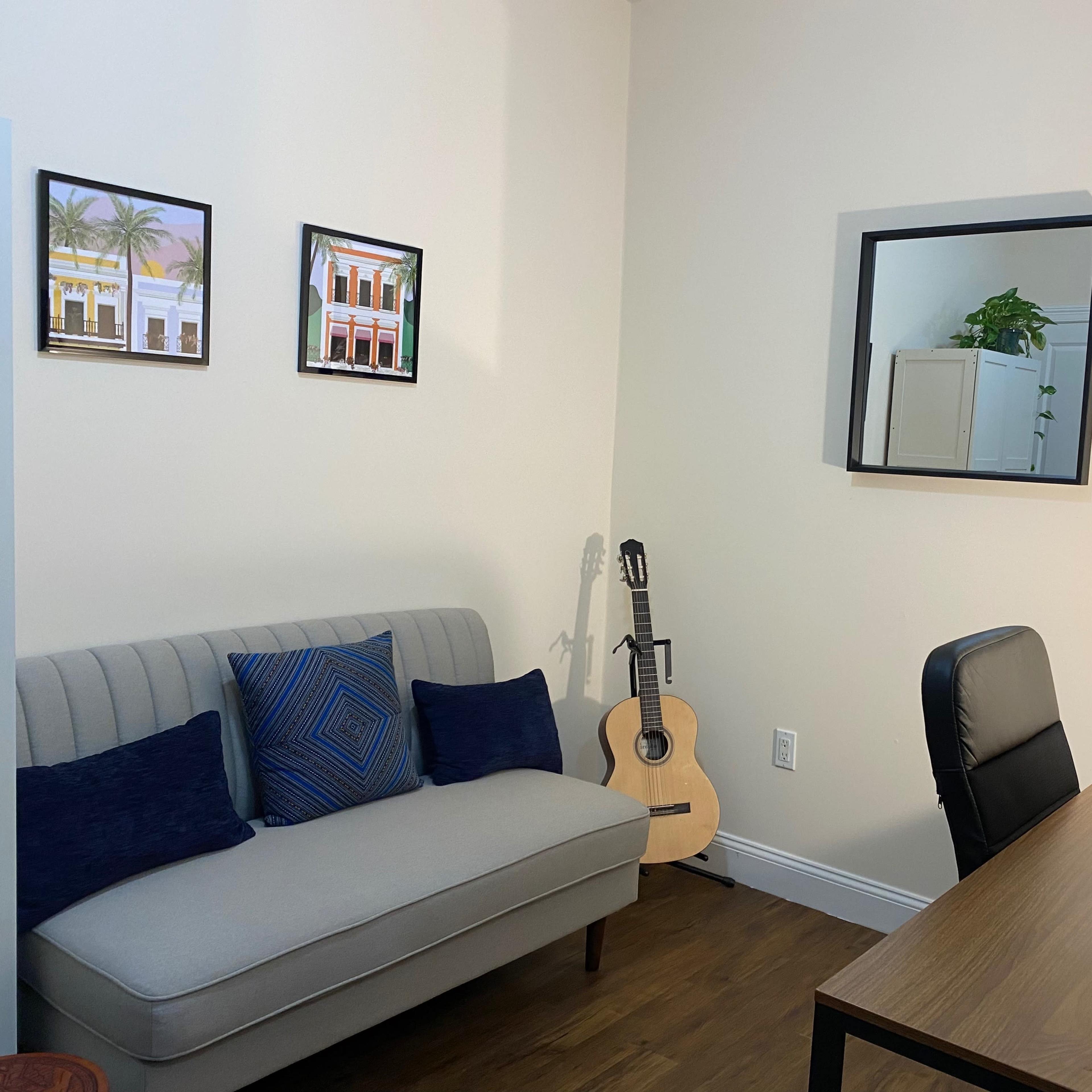 A tidy room with a sofa, a wooden desk with a laptop, a guitar standing against the wall, and framed pictures hanging on the wall.