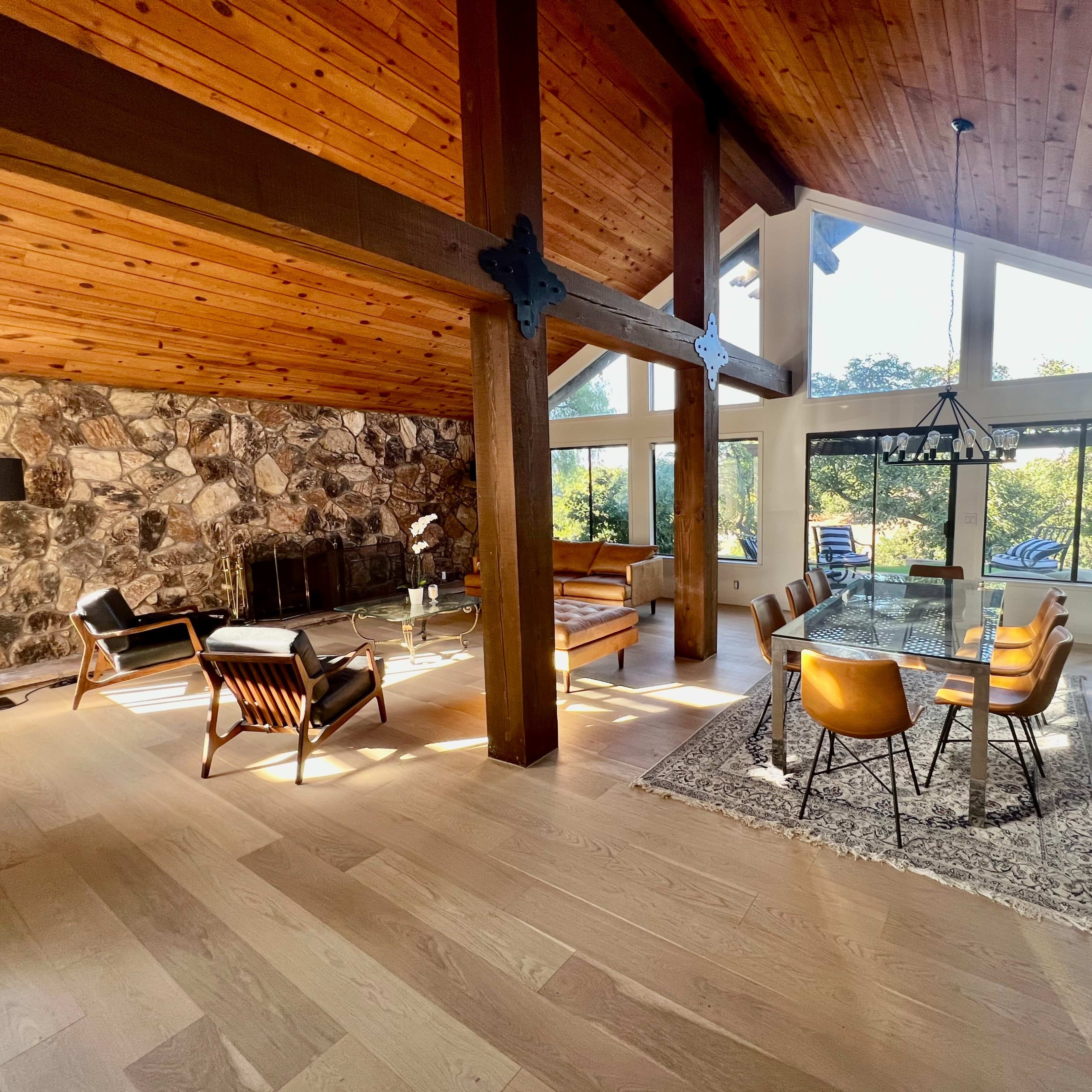 A modern living space with a wooden ceiling, stone wall, and a combination of seating areas and a dining table bathed in natural light from large windows.