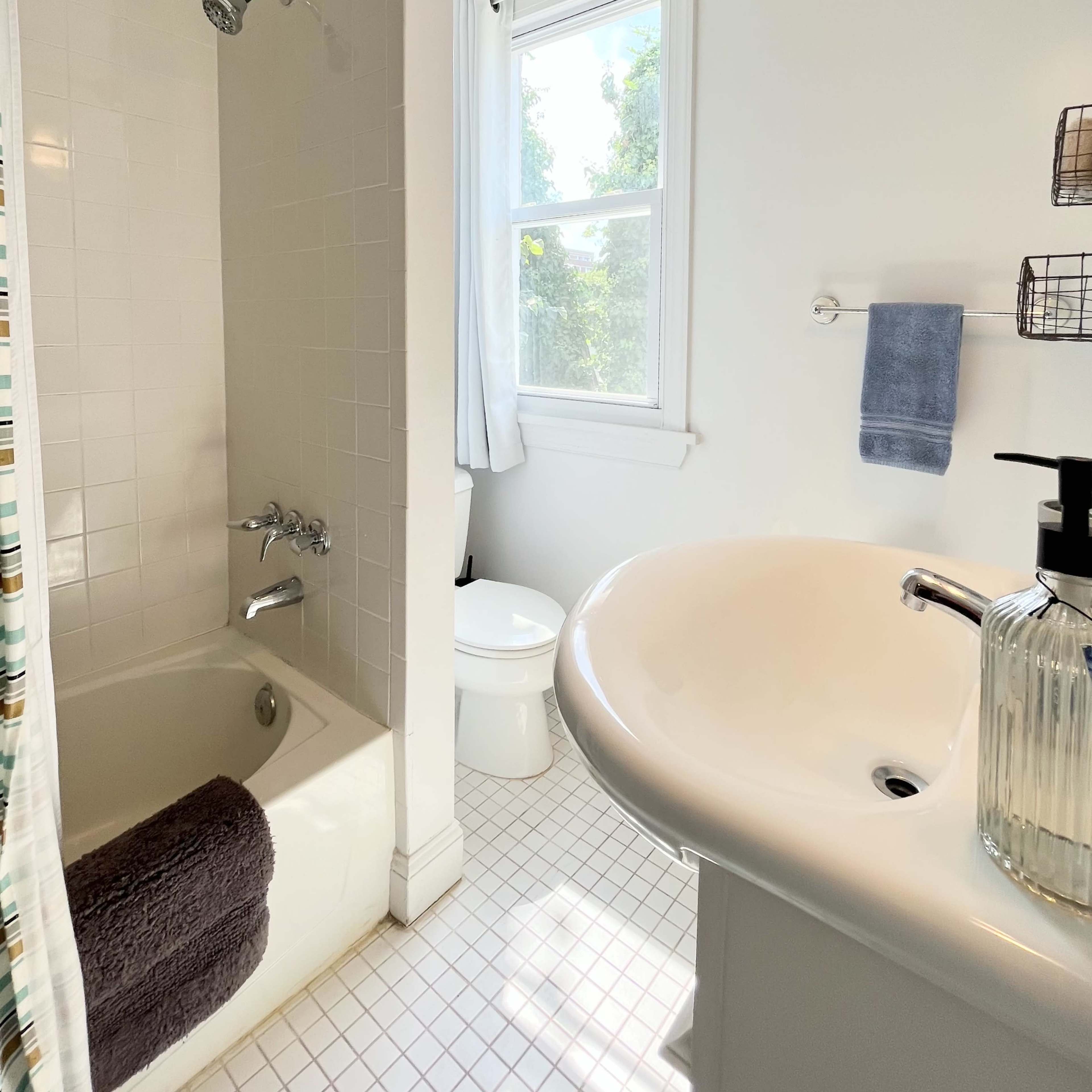 A bathroom featuring a bathtub with a showerhead, a toilet, and a sink with a soap dispenser, illuminated by natural light from a window.