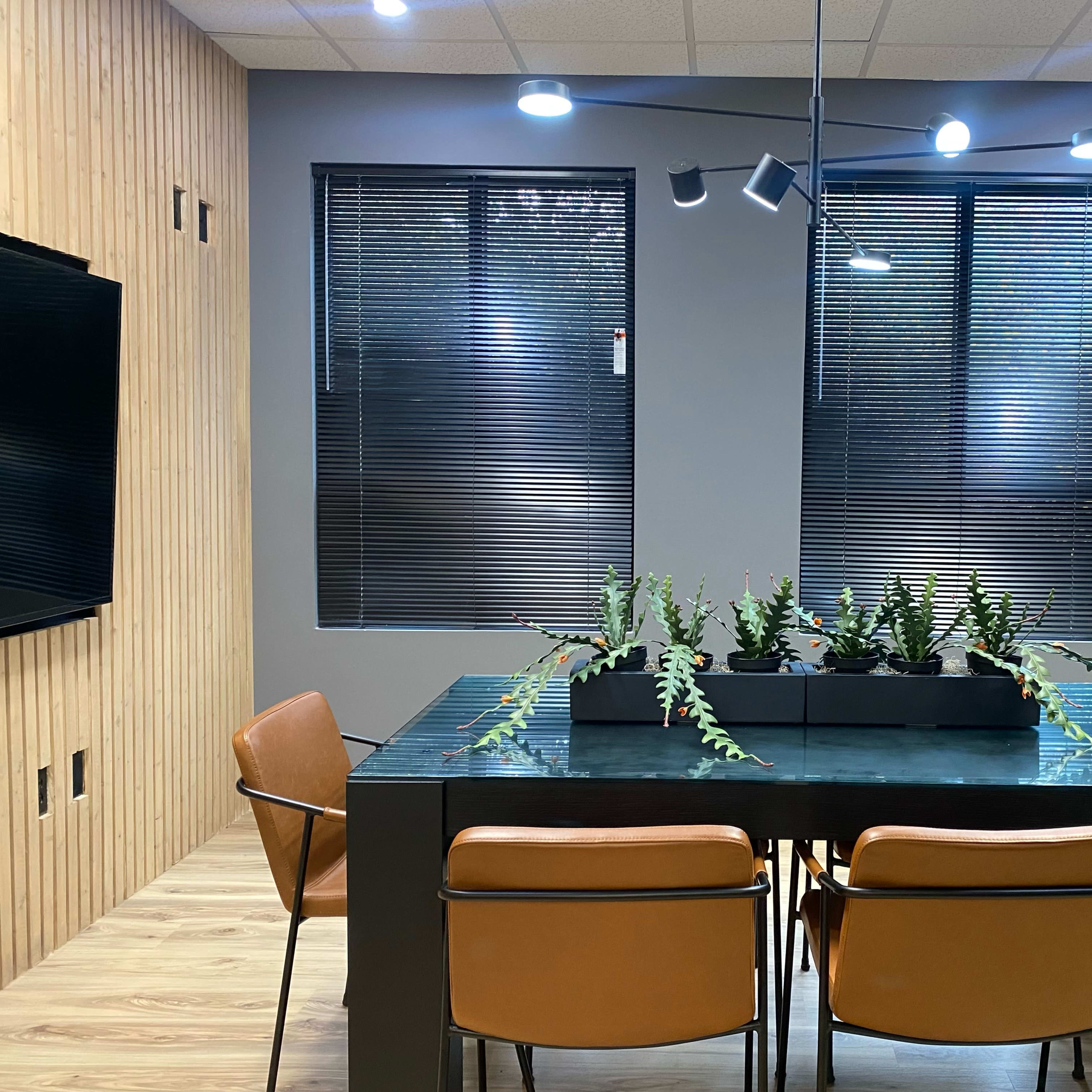 A modern conference room with a glass table surrounded by four brown chairs and a planter of green plants in the center, flanked by windows with blinds.
