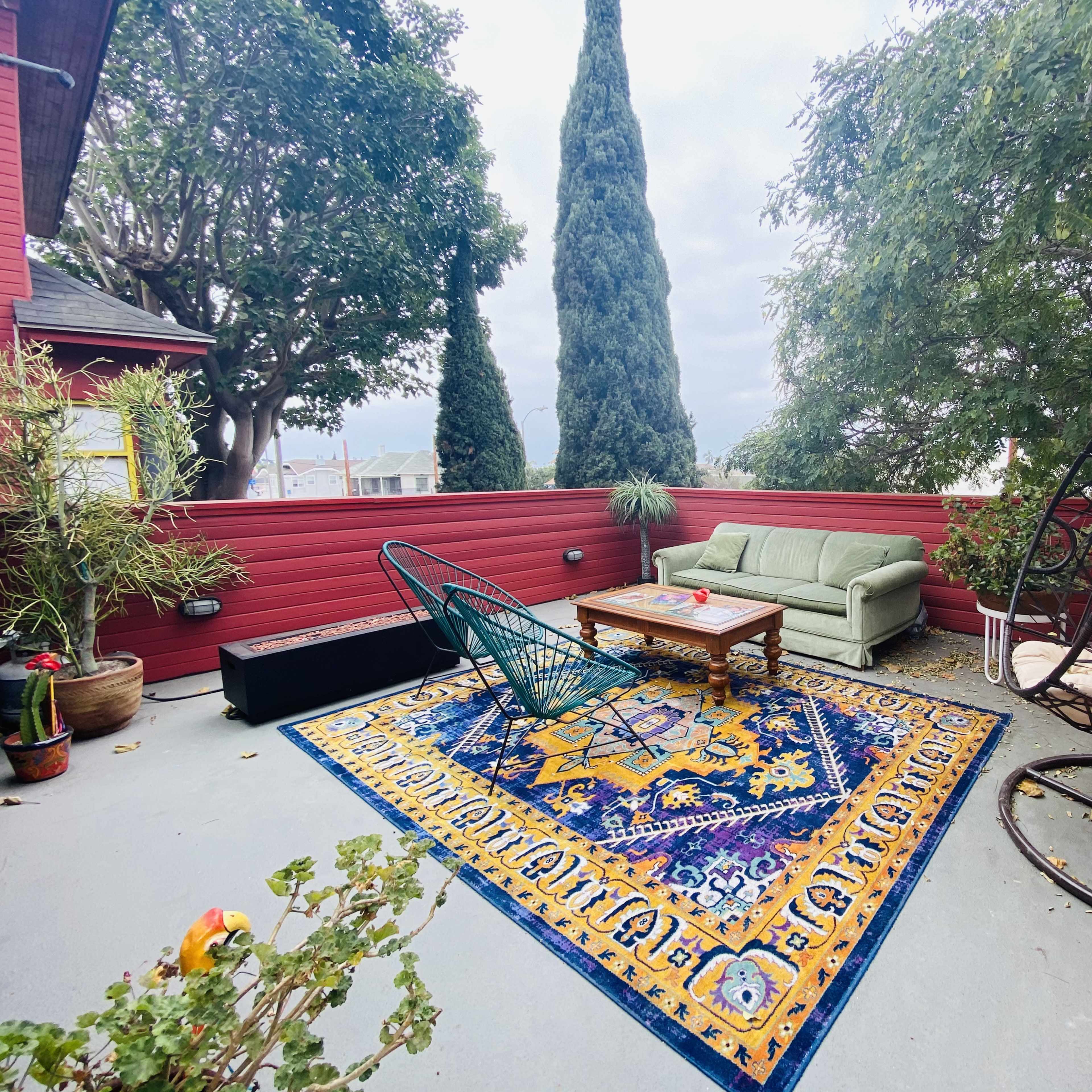 A patio area with a decorative rug, a green sofa, a coffee table, and a hanging chair, surrounded by trees and red wooden walls.