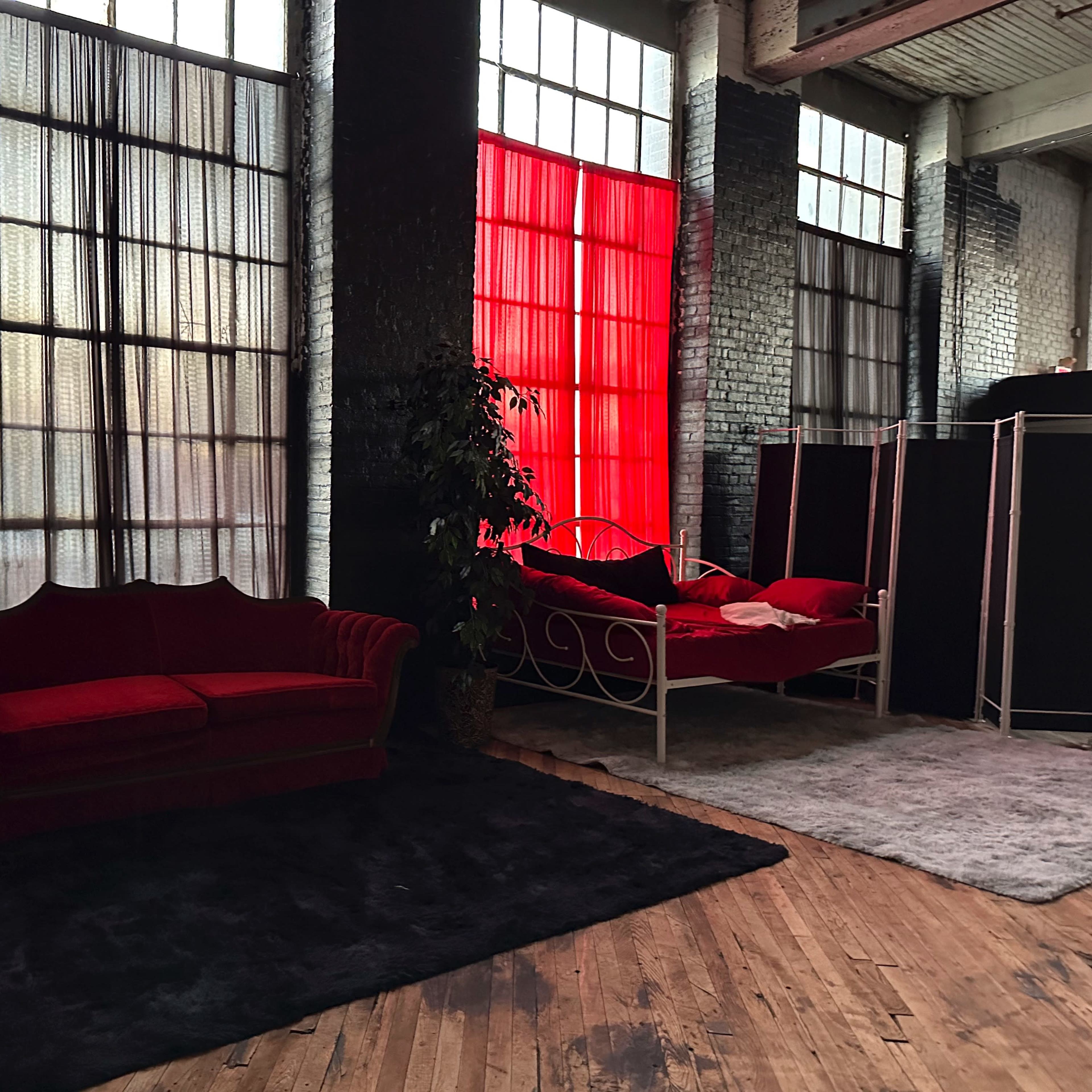 A spacious interior with large windows, featuring a red velvet couch and a white bed against a backdrop of black and red curtains.