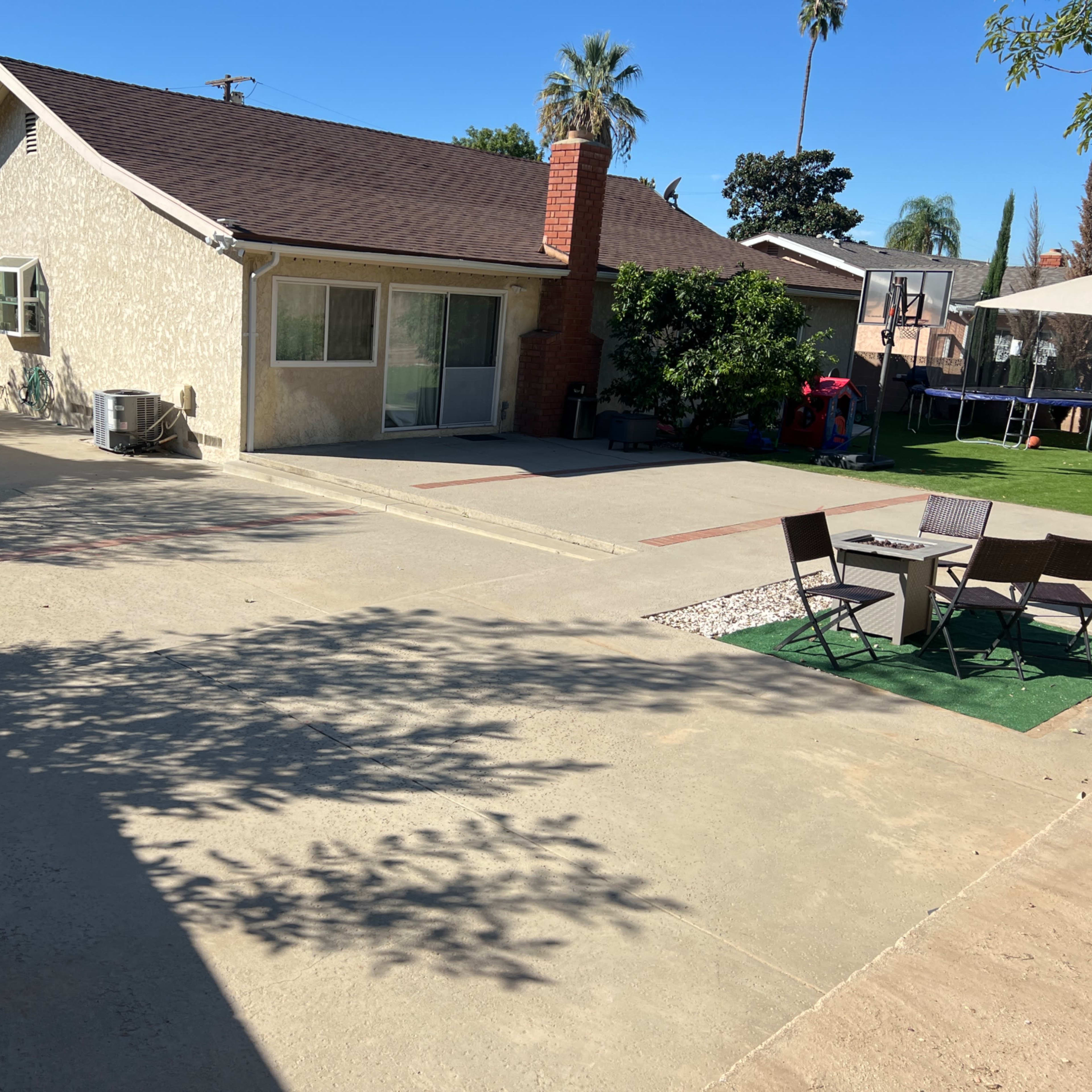 A residential backyard with a concrete patio, a house with a chimney, and outdoor seating arranged around a small grassy area.