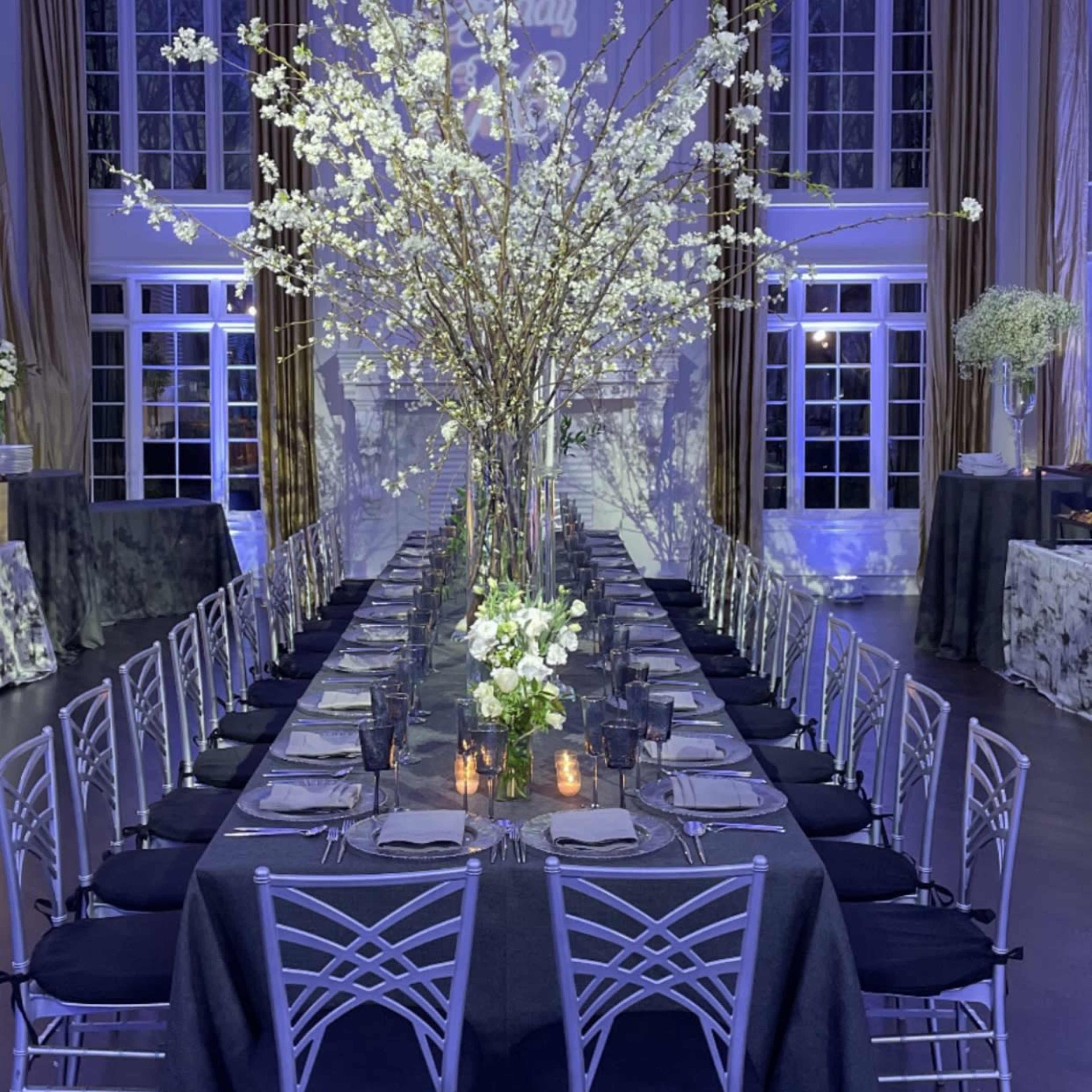 A long dining table is set in a spacious room with elegant decor, featuring tall floral arrangements and a warm glow from lighting.