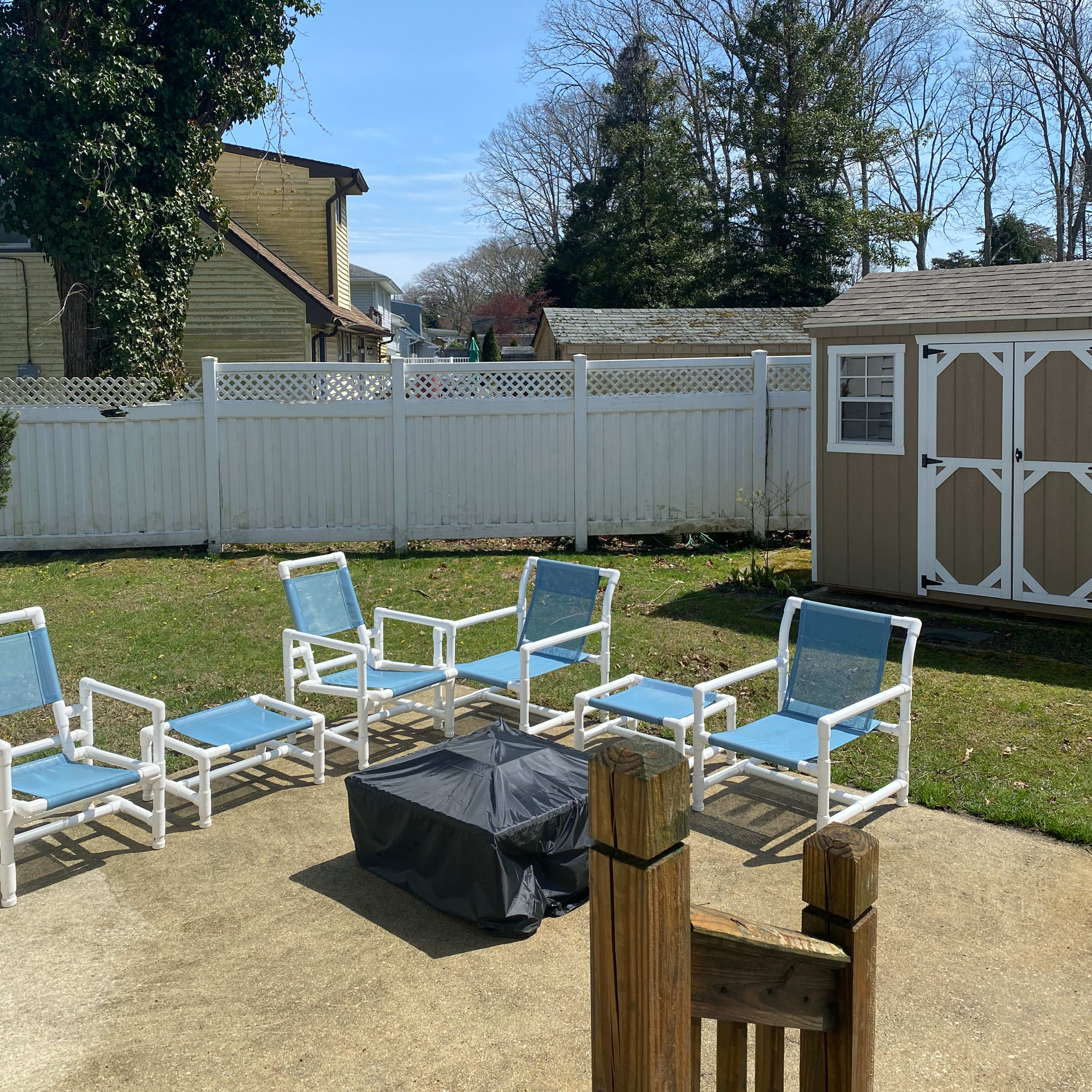 A backyard area featuring six white patio chairs arranged around a black rectangular table, with a shed and a white picket fence in the background under a clear blue sky.