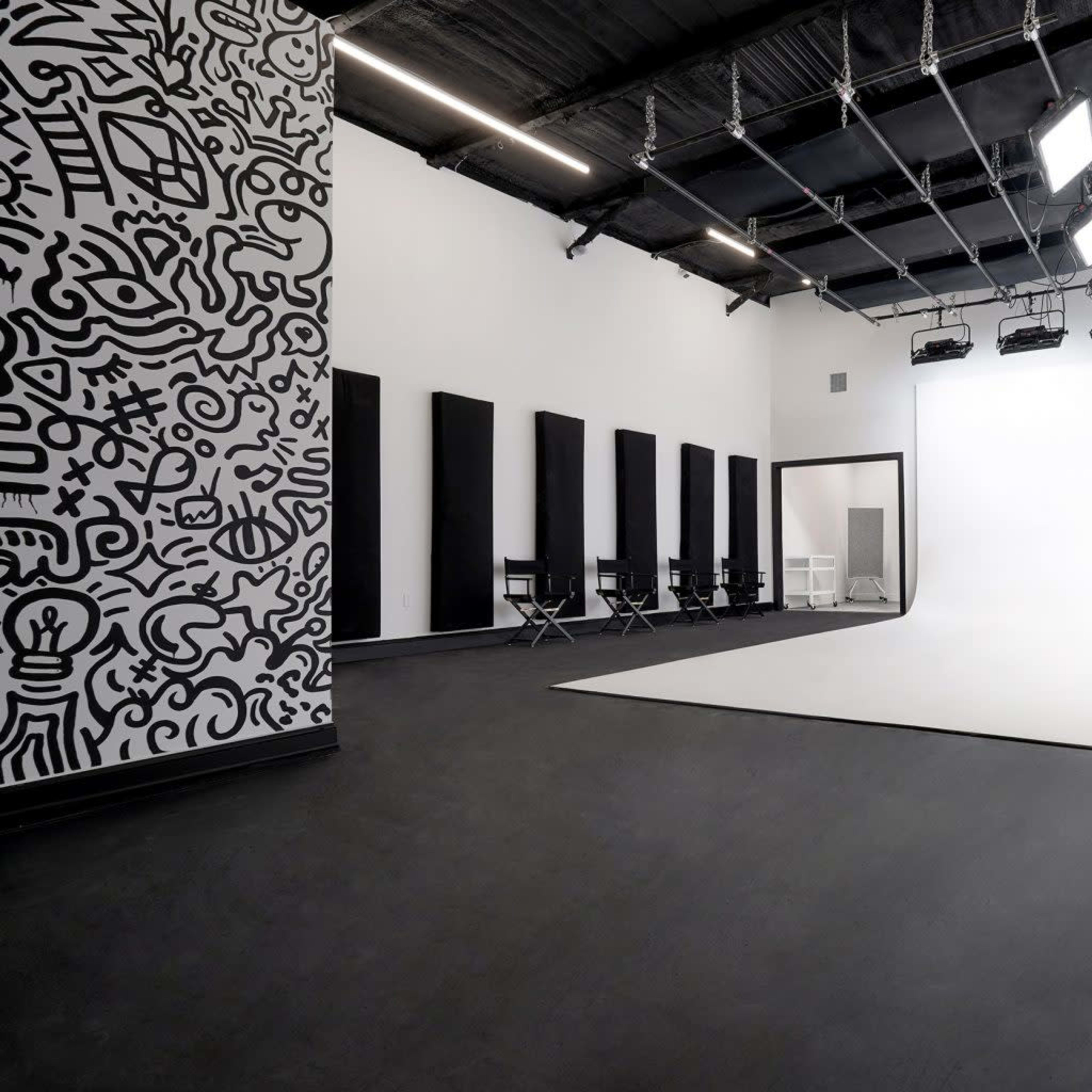 A studio space with a white curved backdrop, black panels on the walls, a patterned mural, and several director's chairs arranged in the corner.