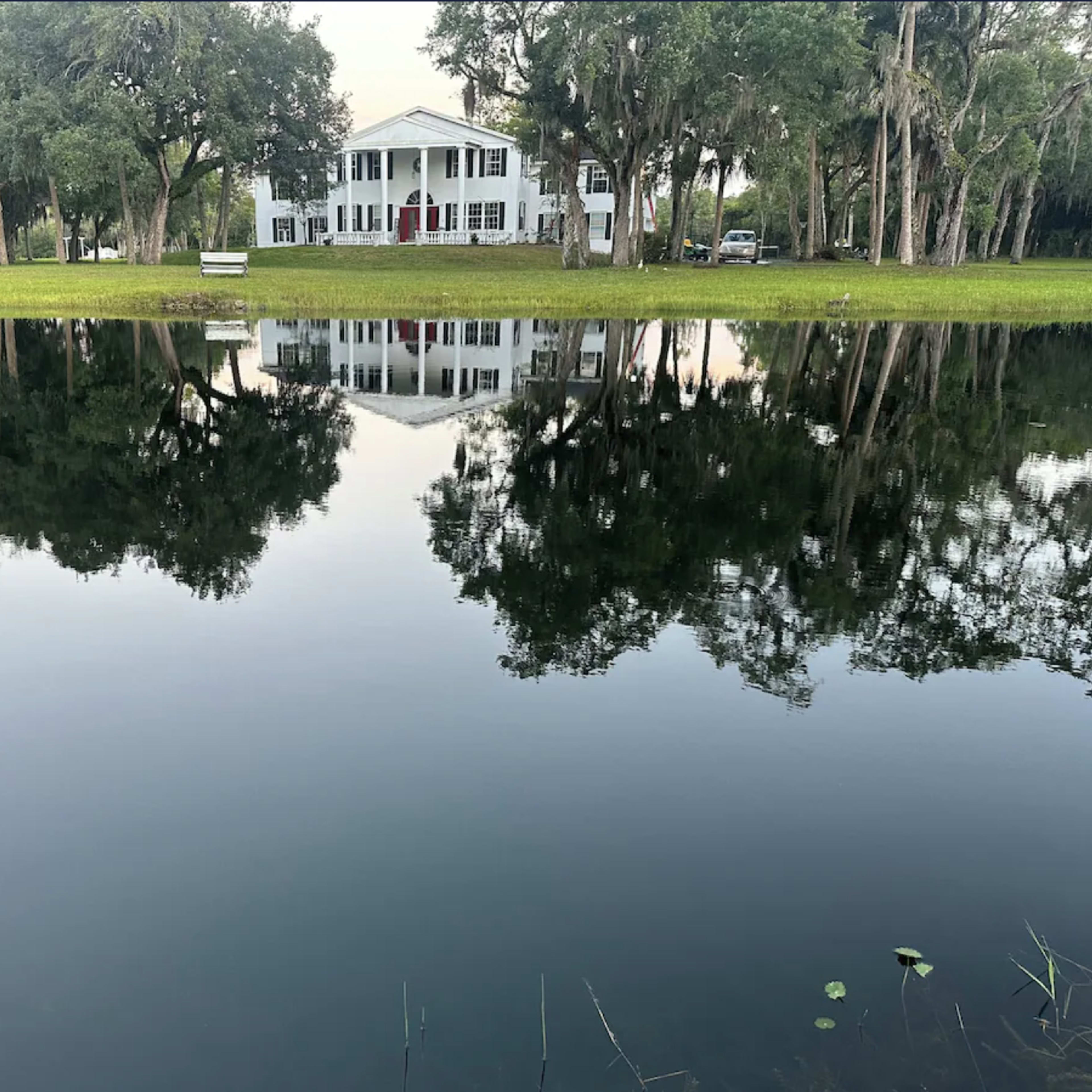 A large white house with a red door stands near a calm pond, reflecting its image and surrounding trees.